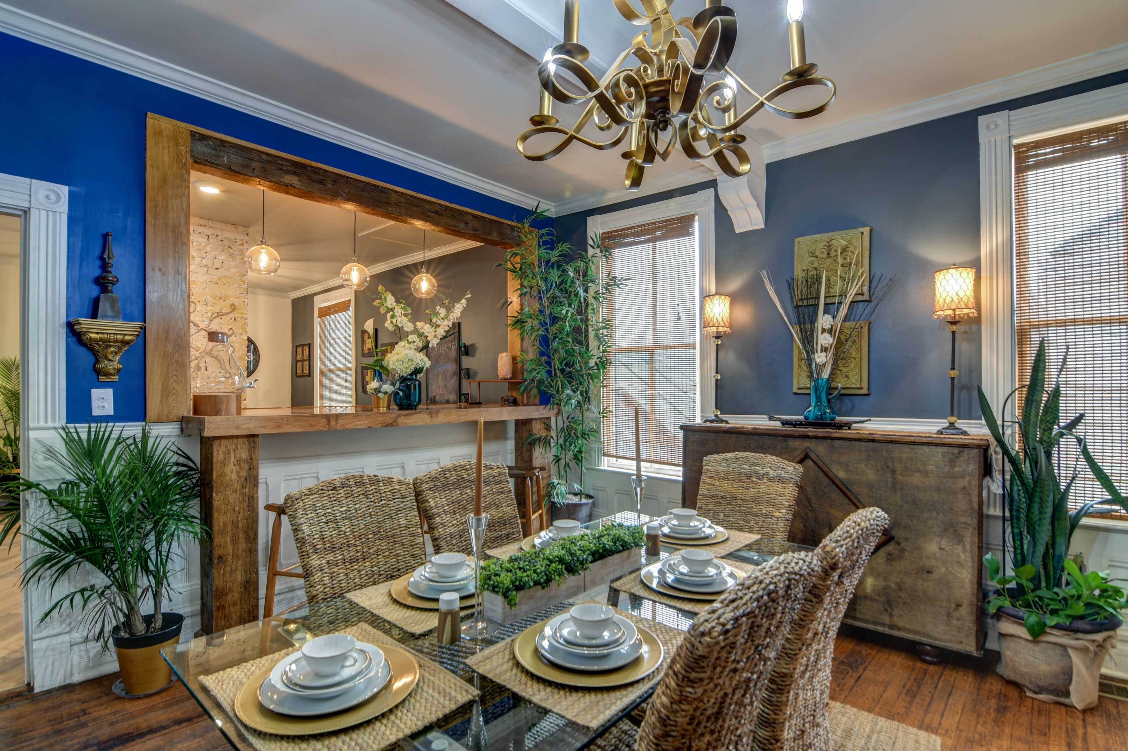 A dining room features a glass table set with plates and a centerpiece, surrounded by woven chairs, with blue walls and a wooden sideboard.