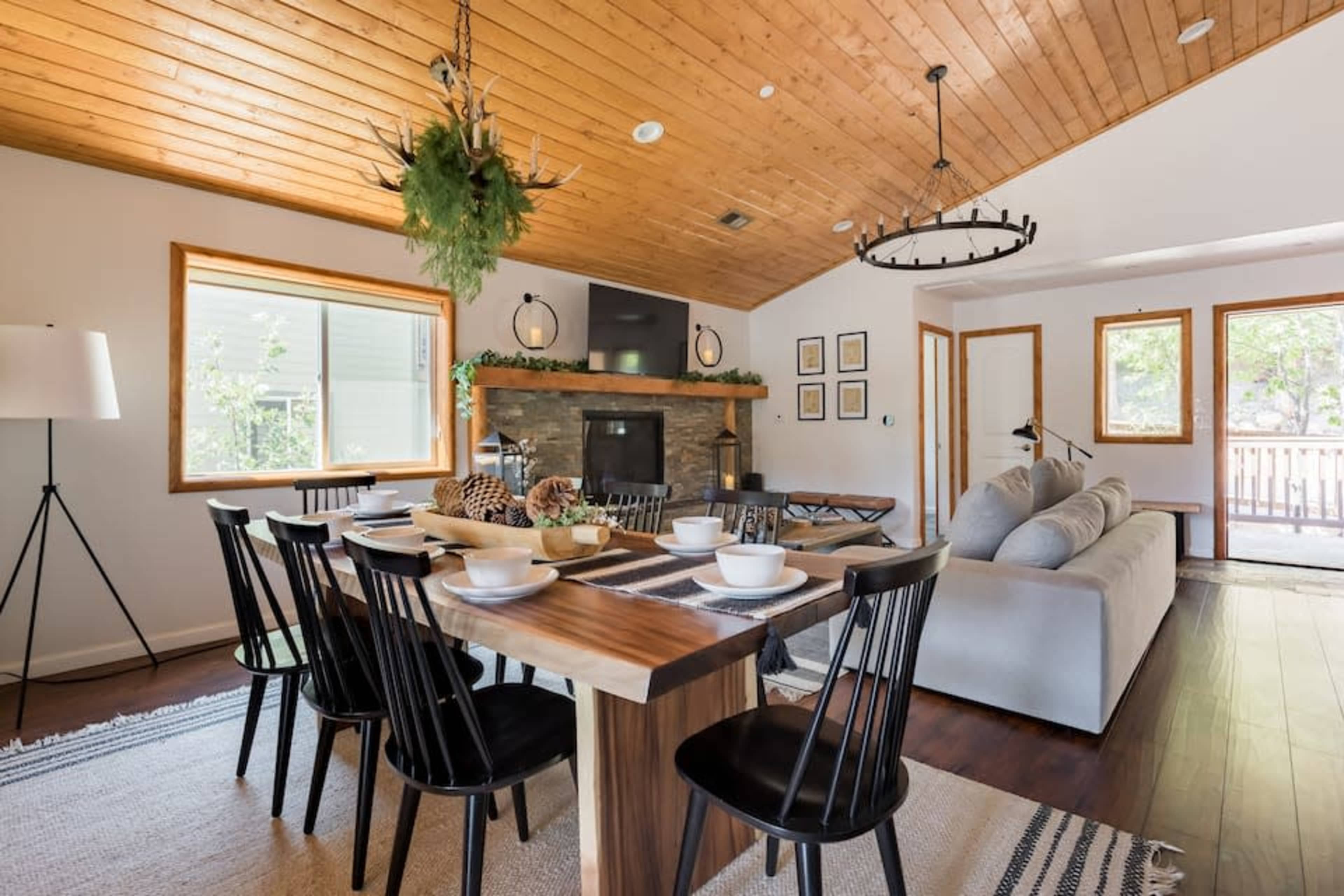 A dining area features a wooden table set for meals, surrounded by black chairs, while a living space with a sofa, a stone fireplace, and large windows is visible in the background.