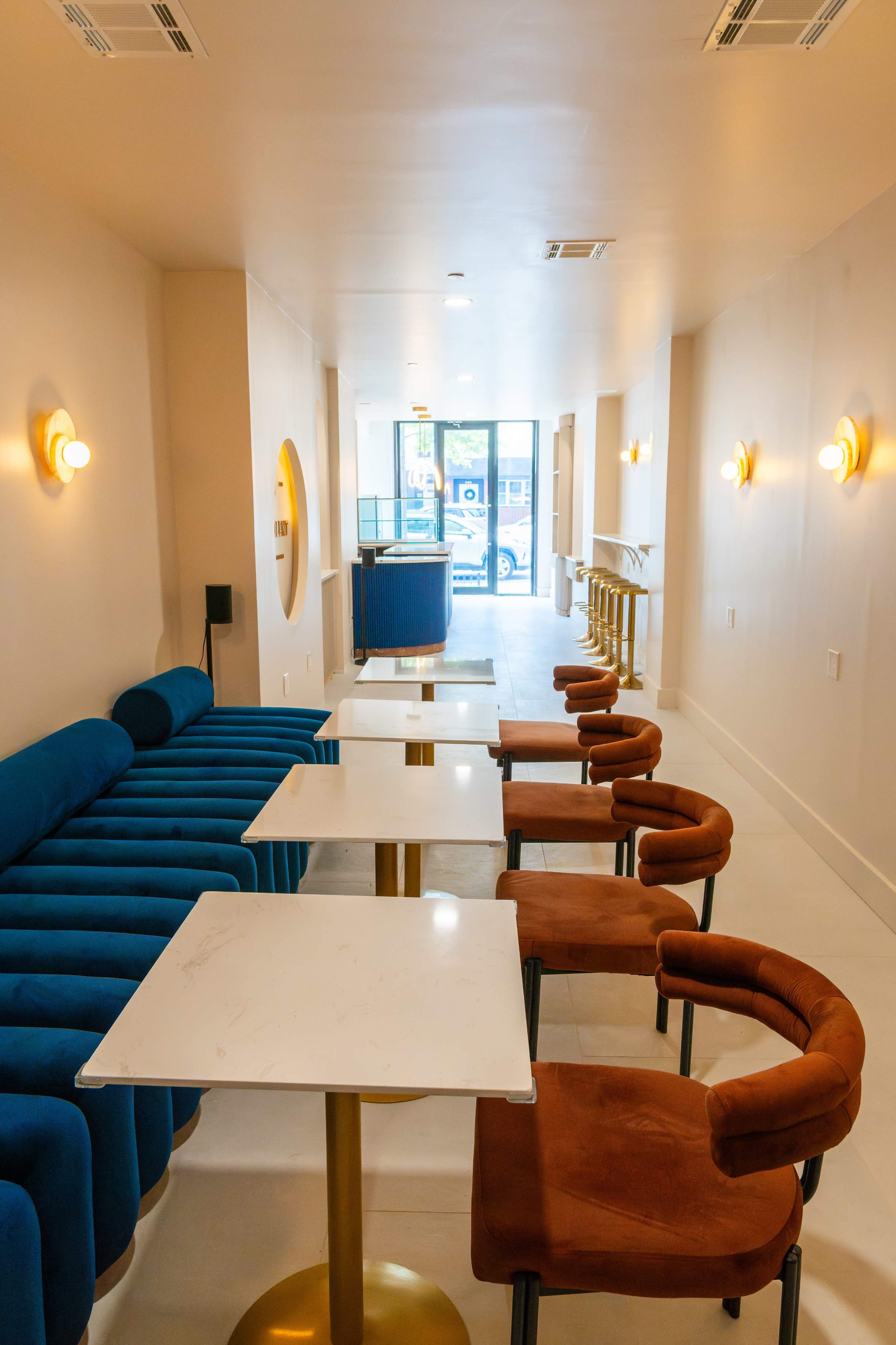 The image shows a modern cafe interior with blue sofas, brown upholstered chairs, and white marble tables lined up in a well-lit area.