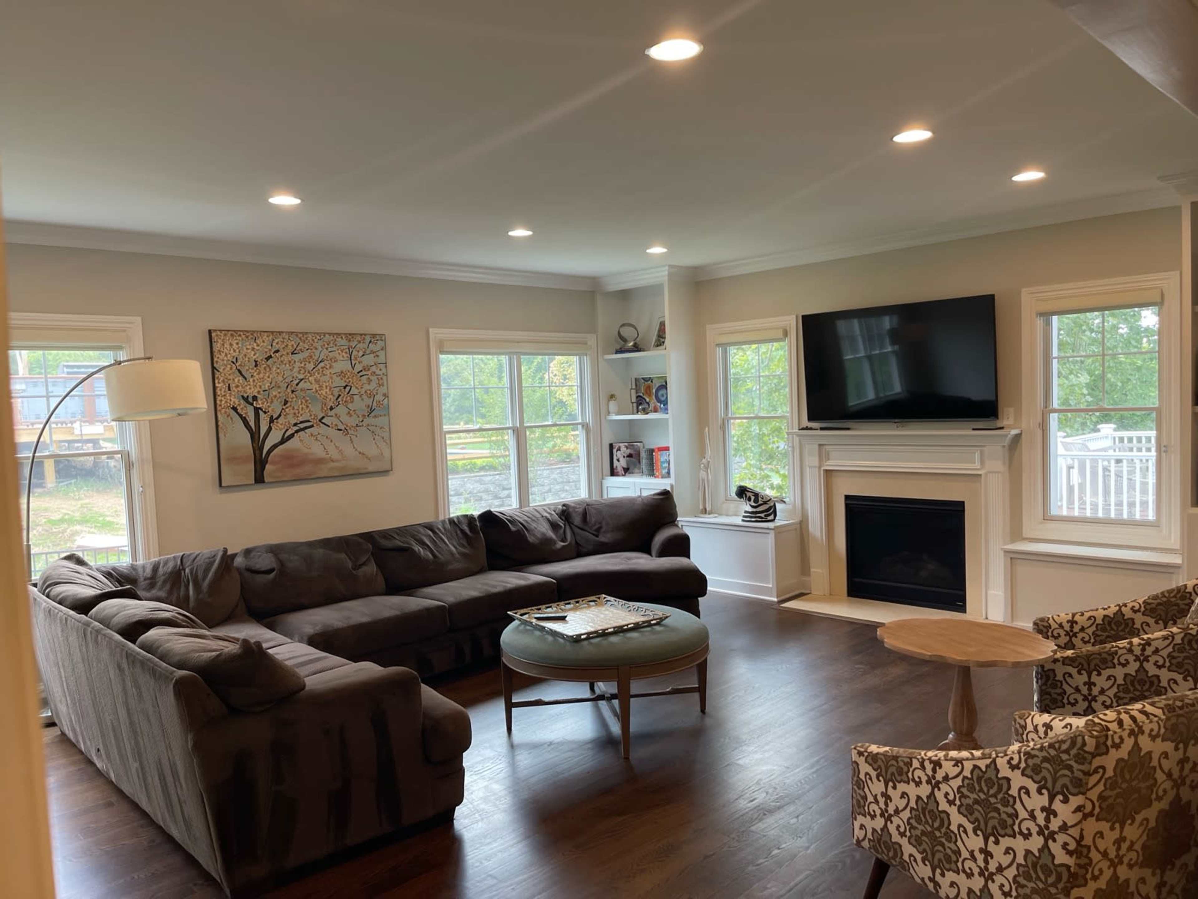 The image shows a living room with a large sectional sofa, a round coffee table, a flat-screen television mounted above a fireplace, and large windows that let in natural light.