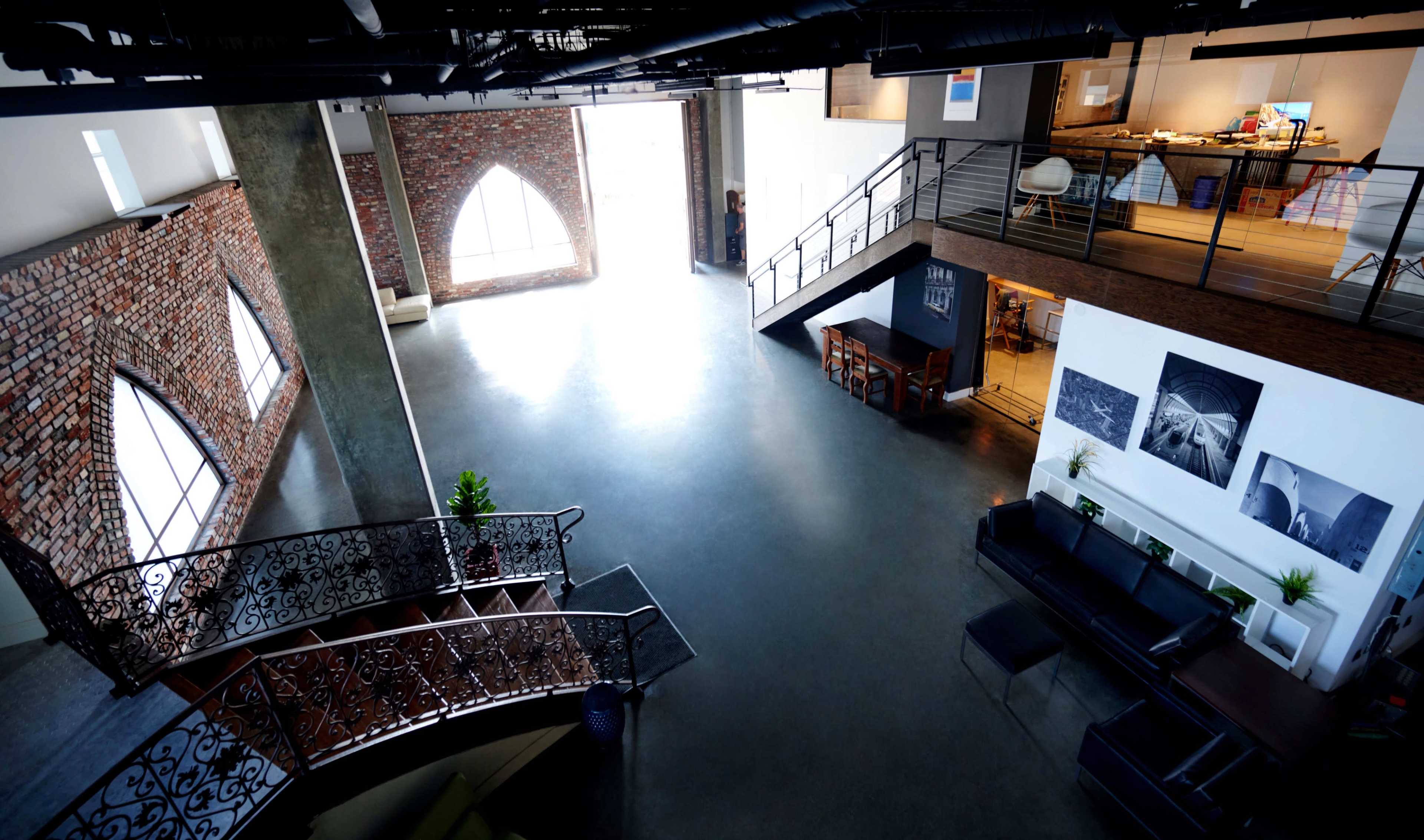 The image shows a spacious, modern interior of a building featuring a wooden staircase, exposed brick walls, and a large open area with seating.
