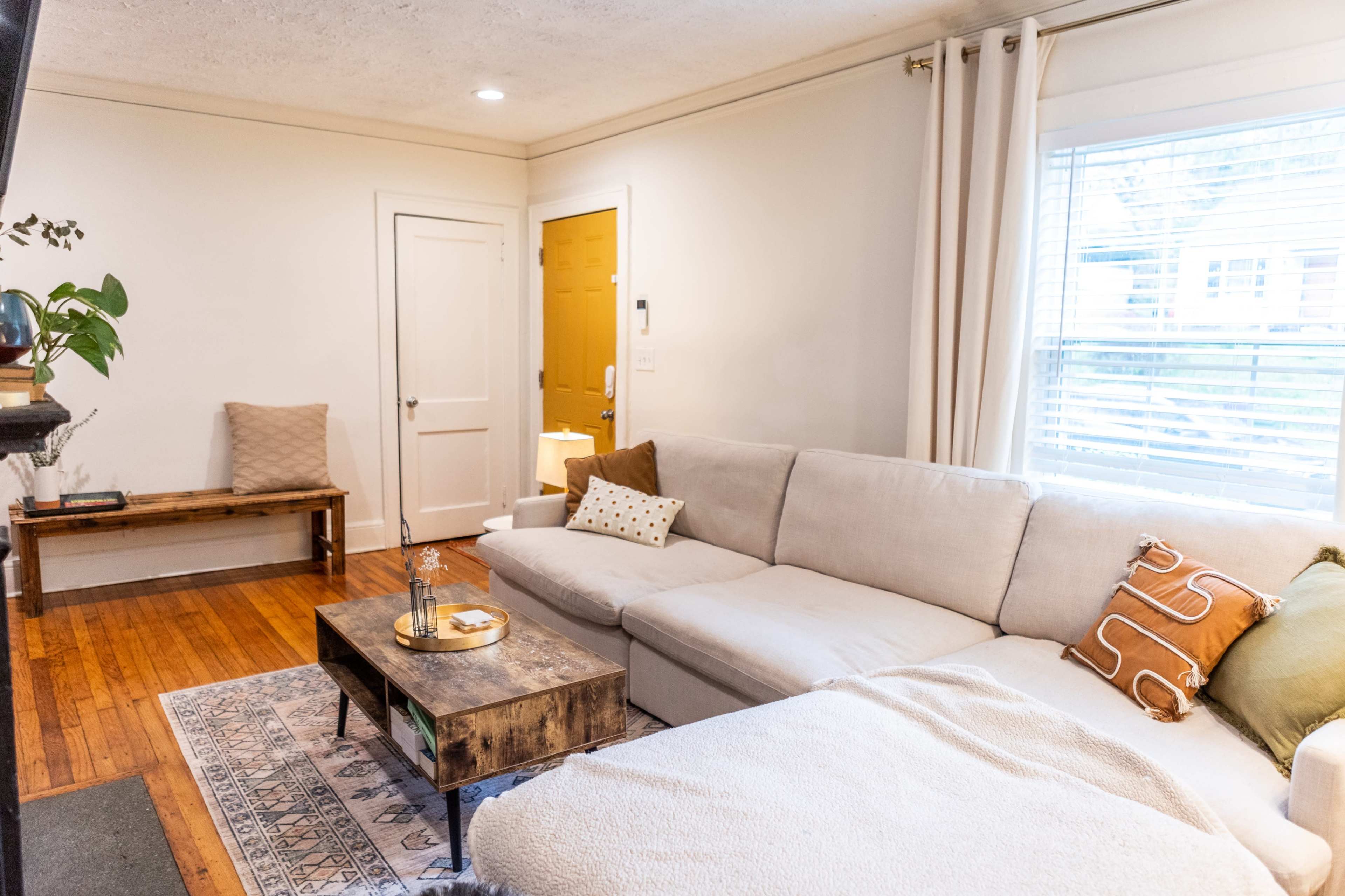 The image shows a living room with a sectional sofa, a coffee table, and a door leading outside, illuminated by natural light through a window.
