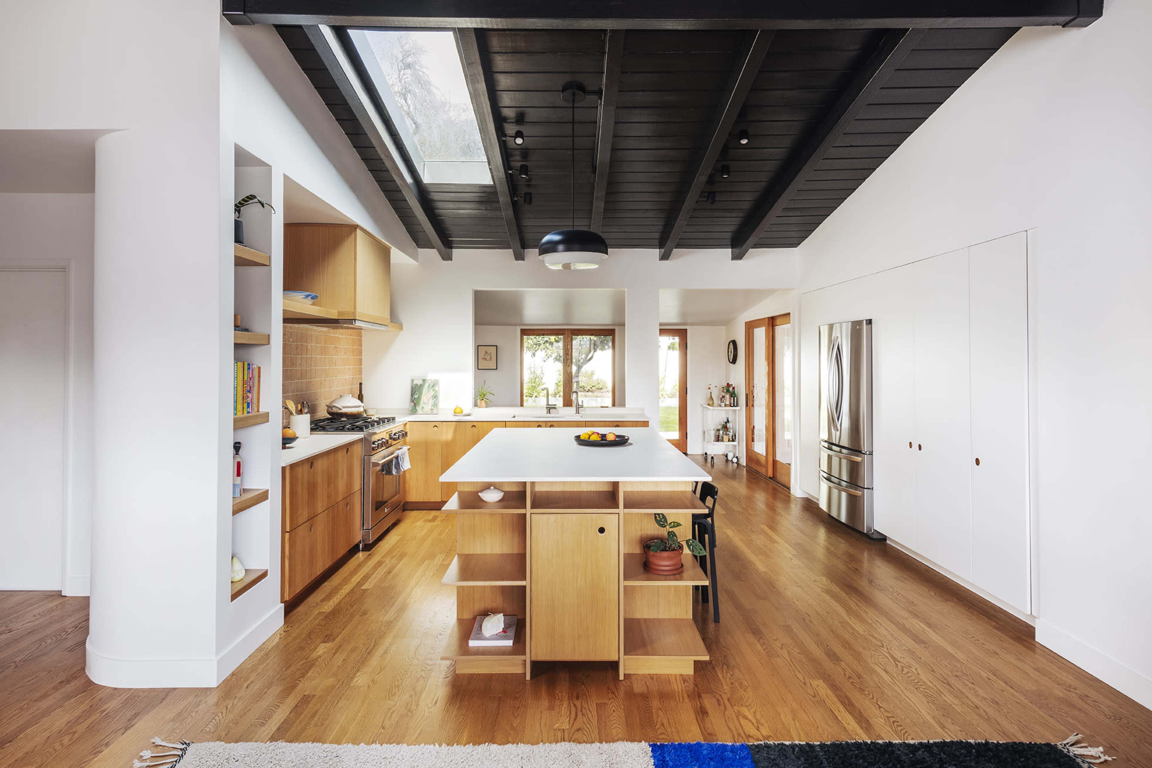 The image shows a modern kitchen featuring wooden cabinetry, an island with open shelving, and large windows that provide natural light.