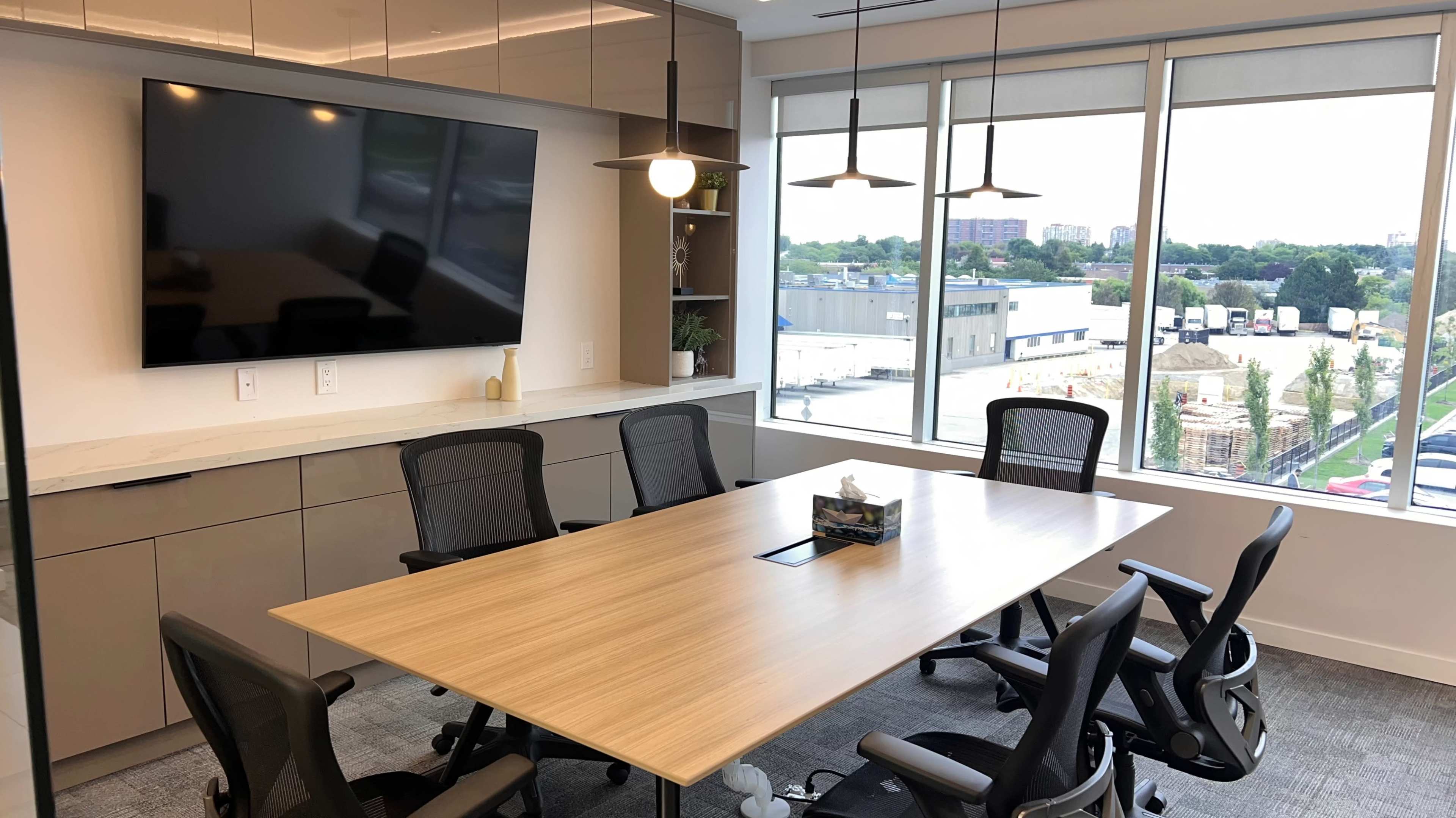 The image shows a modern conference room with a large table, six ergonomic chairs, a wall-mounted television, and a window overlooking an outdoor area with vehicles.