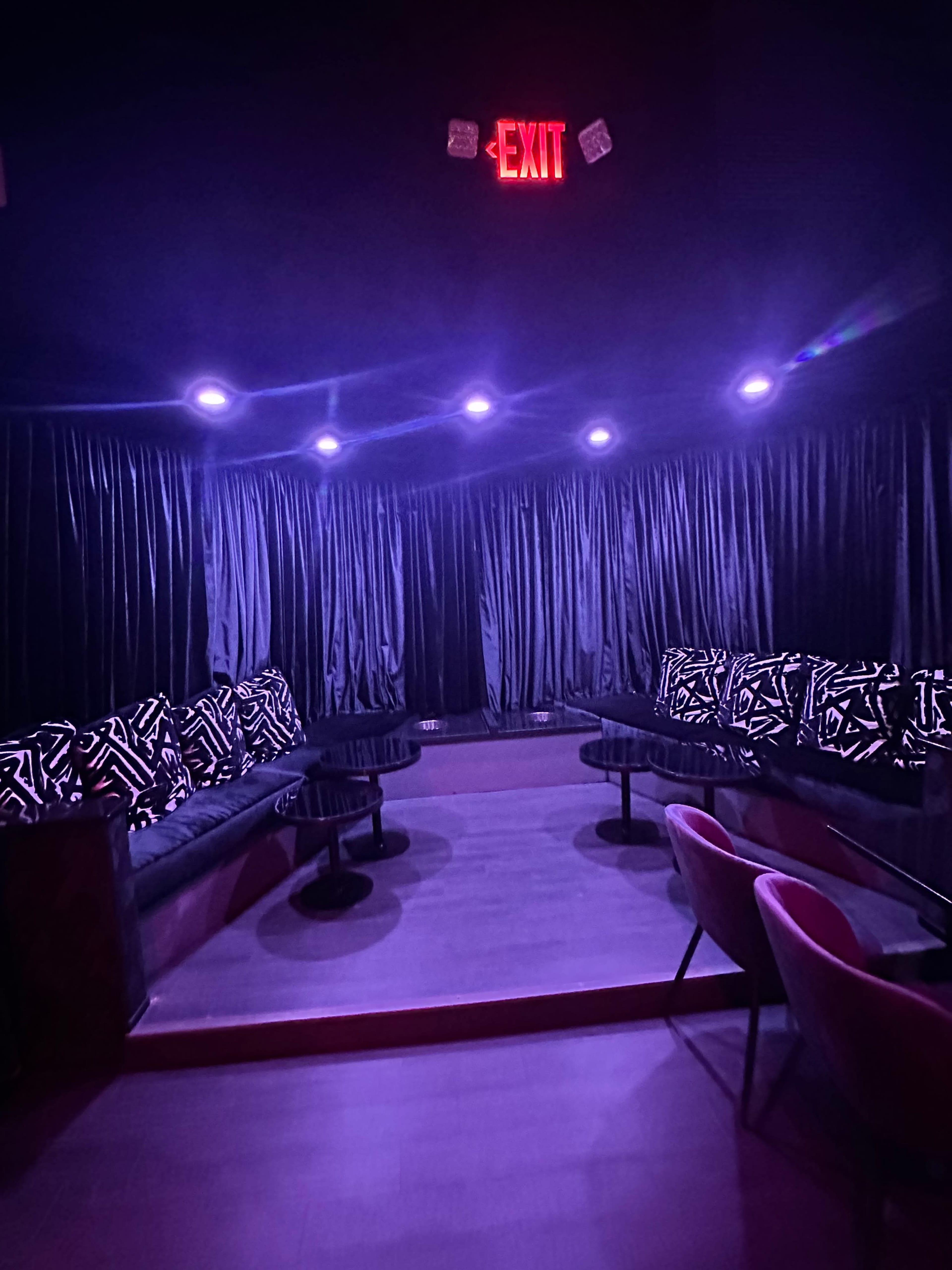 A dimly lit lounge area with dark curtains, patterned cushions on sofas, and an illuminated exit sign above.