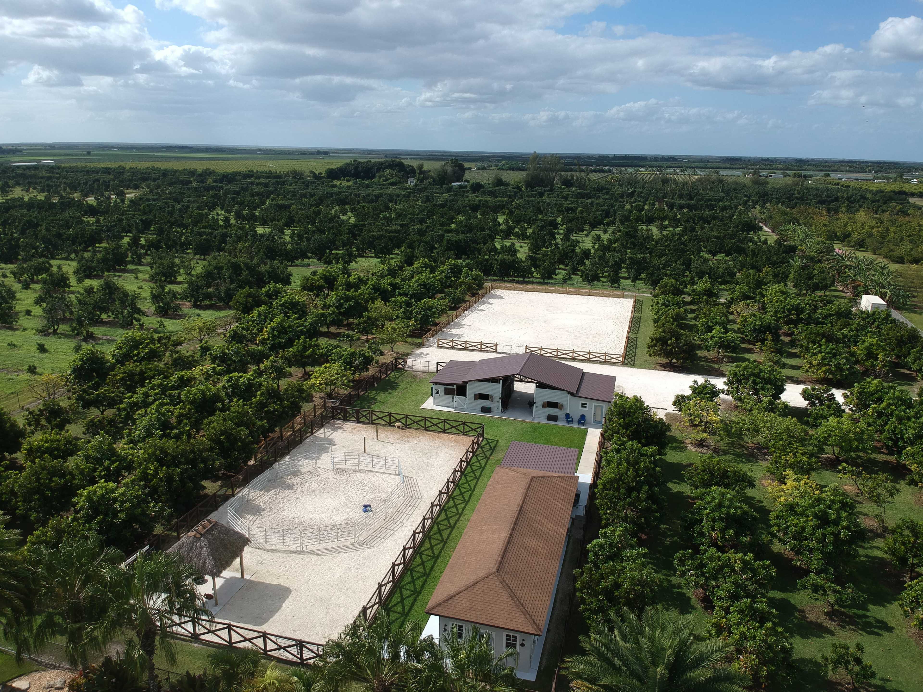 An aerial view shows a property with two fenced riding arenas, a main building, and surrounding greenery.