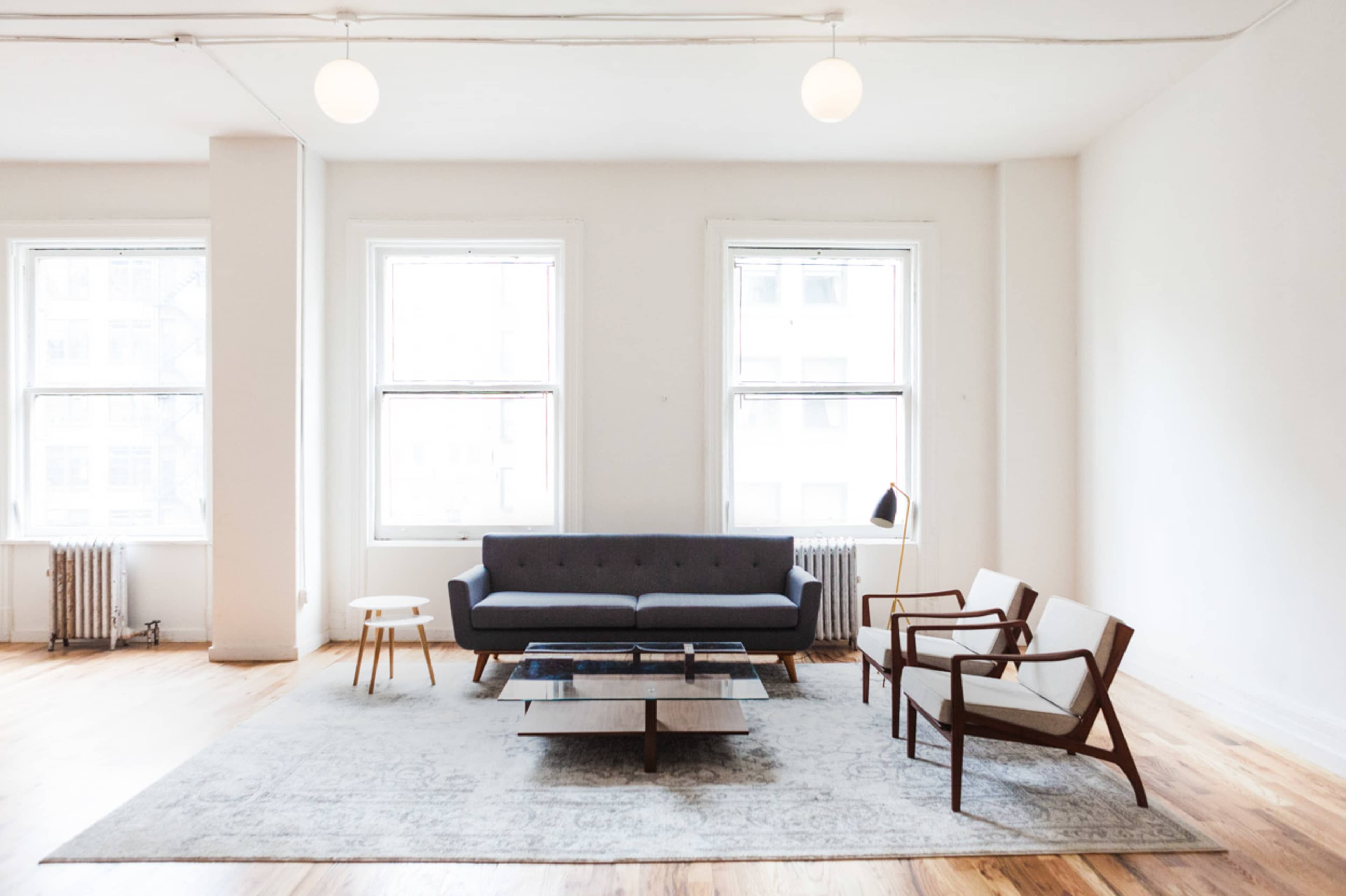 A minimalist living room features a gray sofa, two wooden armchairs, a coffee table, and large windows allowing natural light to fill the space.
