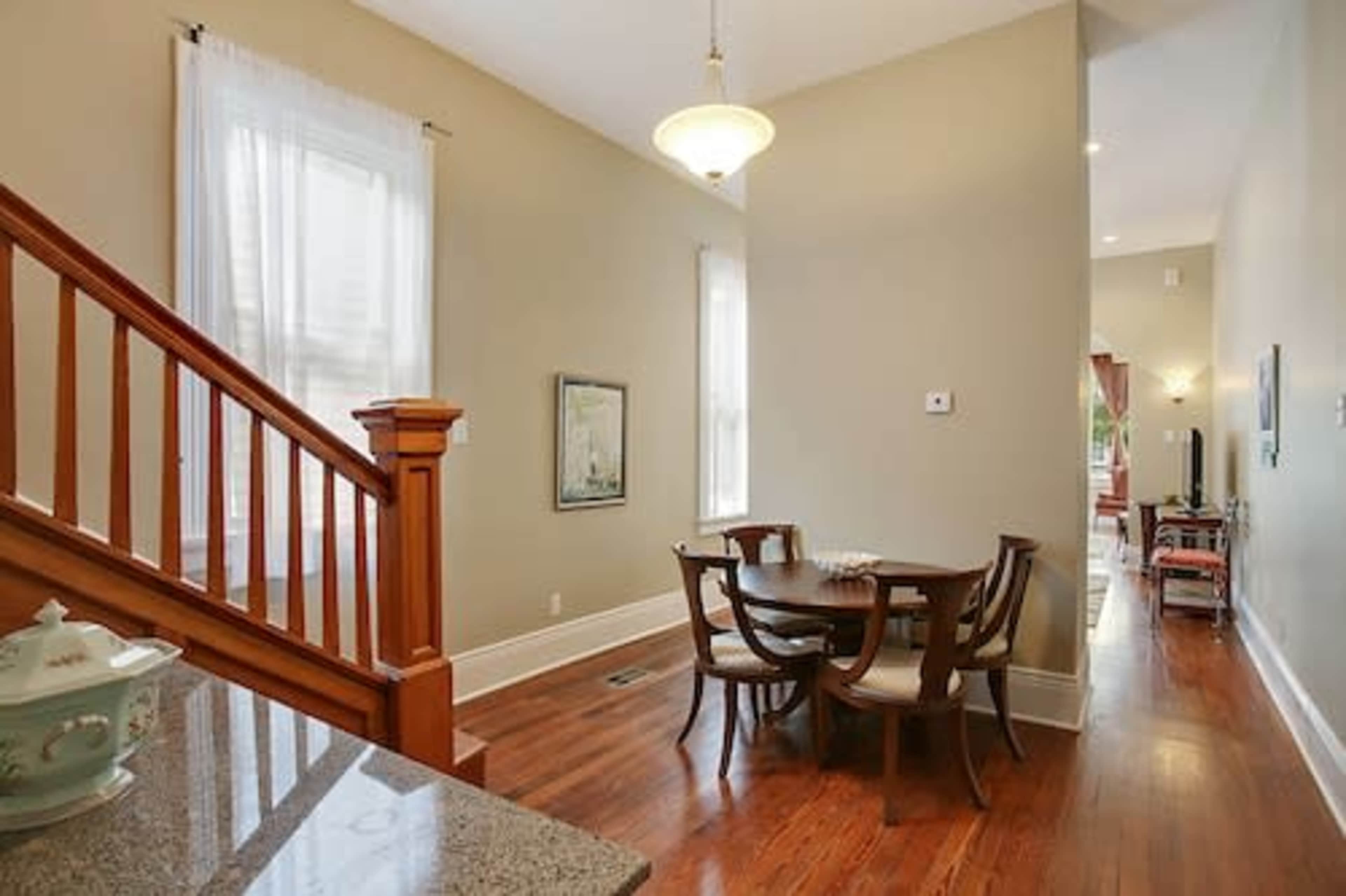 A well-lit dining area featuring a round wooden table surrounded by six chairs, adjacent to a staircase with wooden railings.