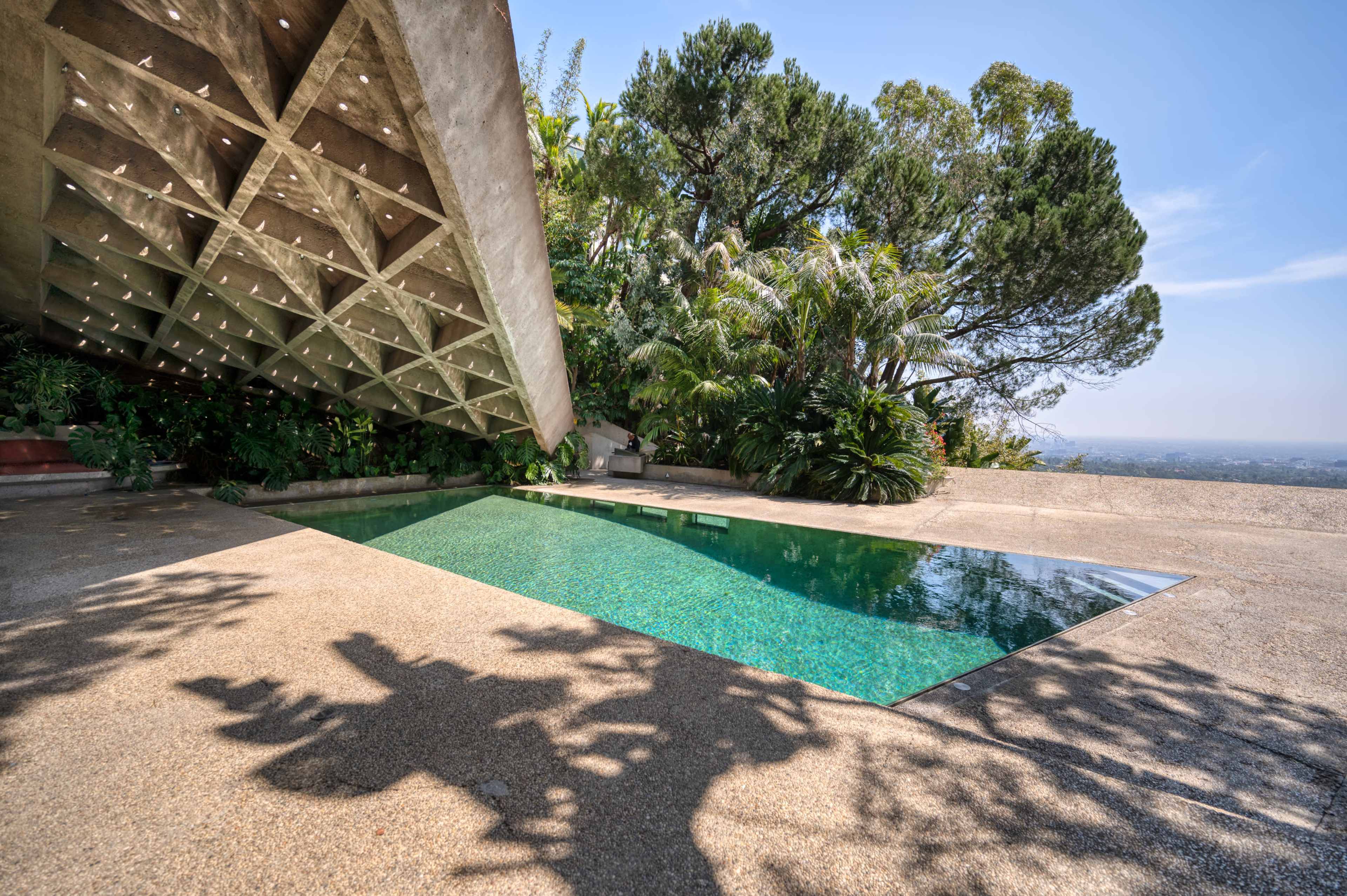 The image shows a modern architectural structure with a slanted roof overlooking a rectangular pool surrounded by lush greenery.