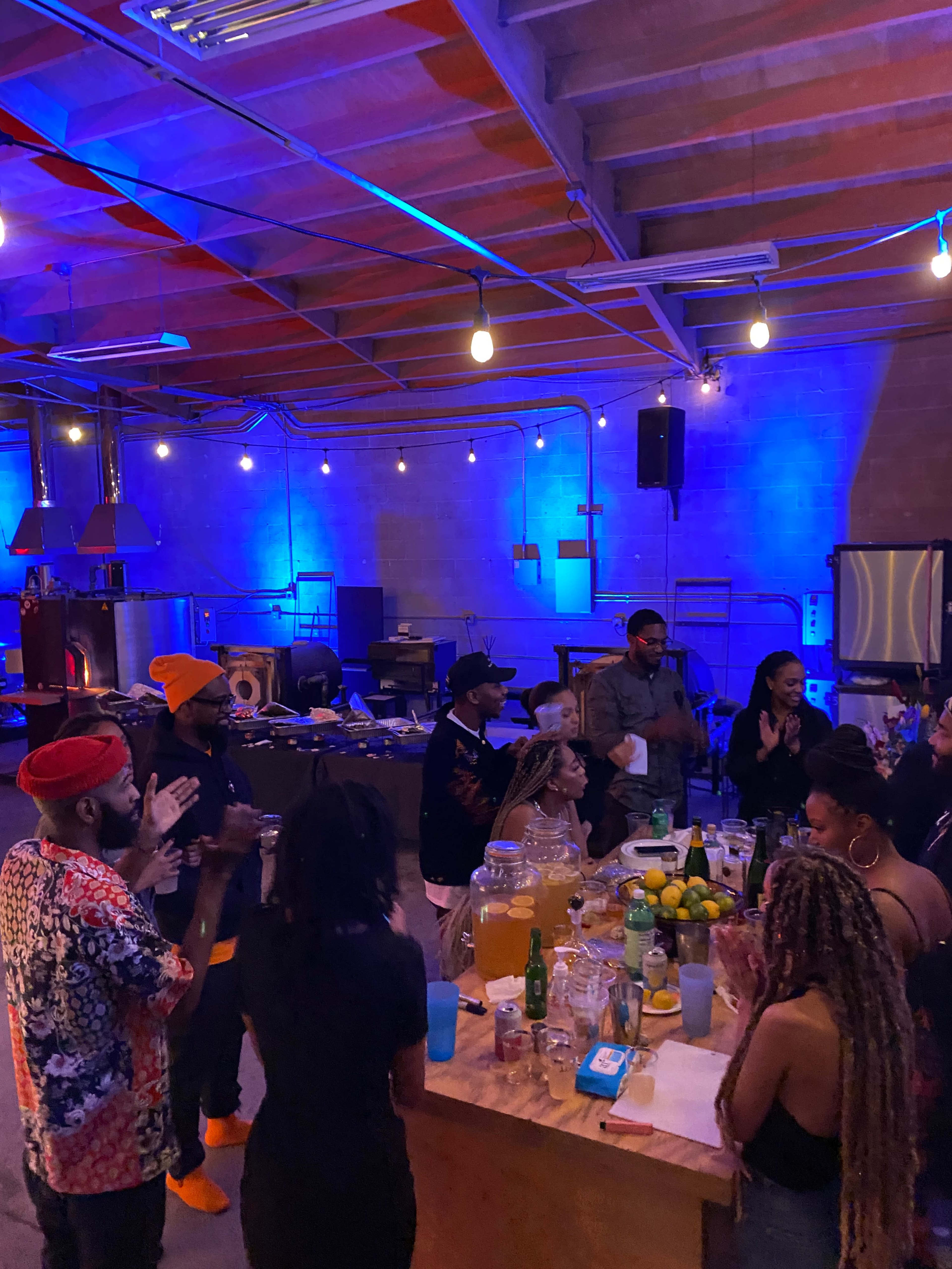 A group of people gathers around a large table filled with drinks and food in a stylish, well-lit space with industrial decor.