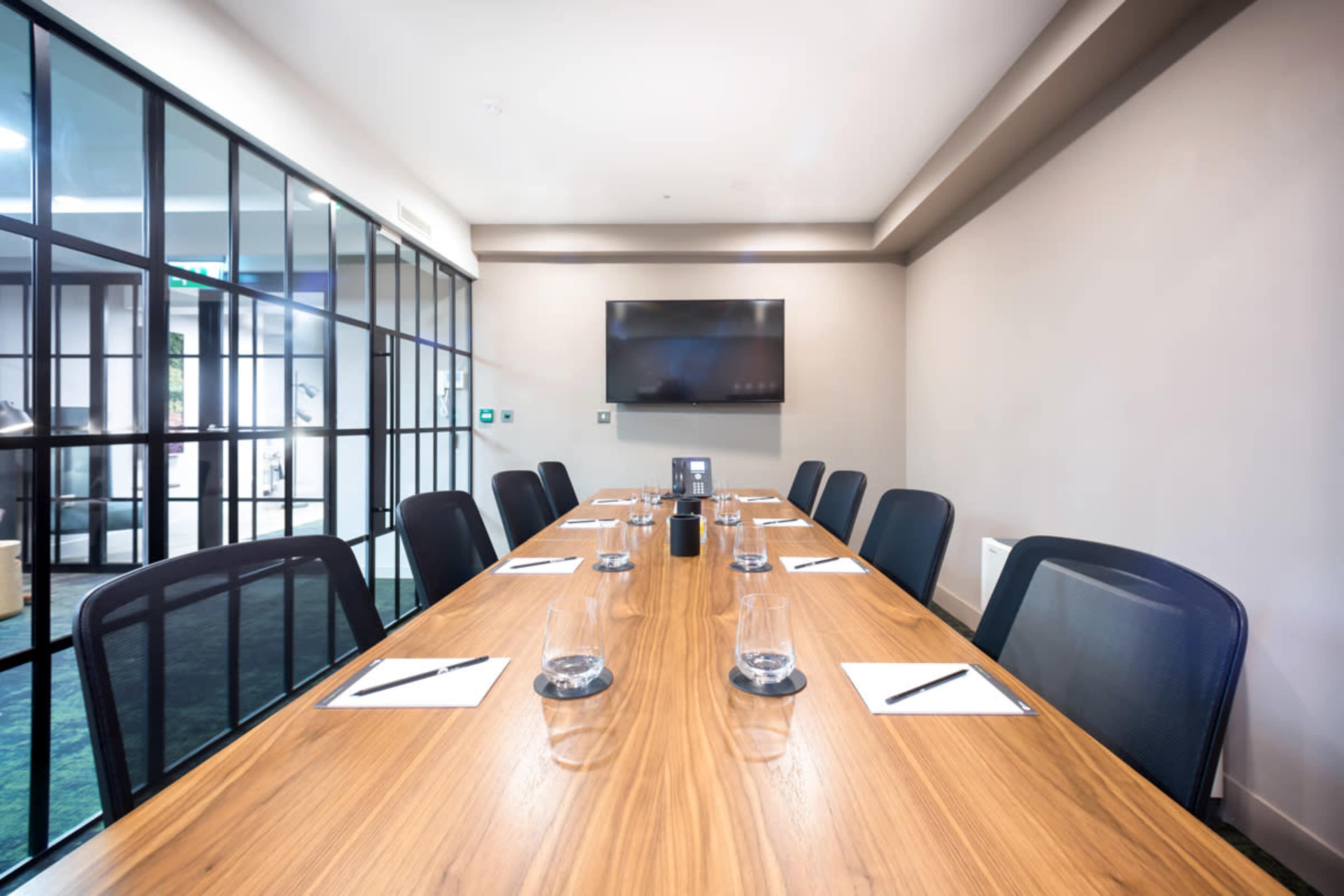 A conference room with a long wooden table set for a meeting, complete with notebooks, pens, and glasses of water, is displayed.