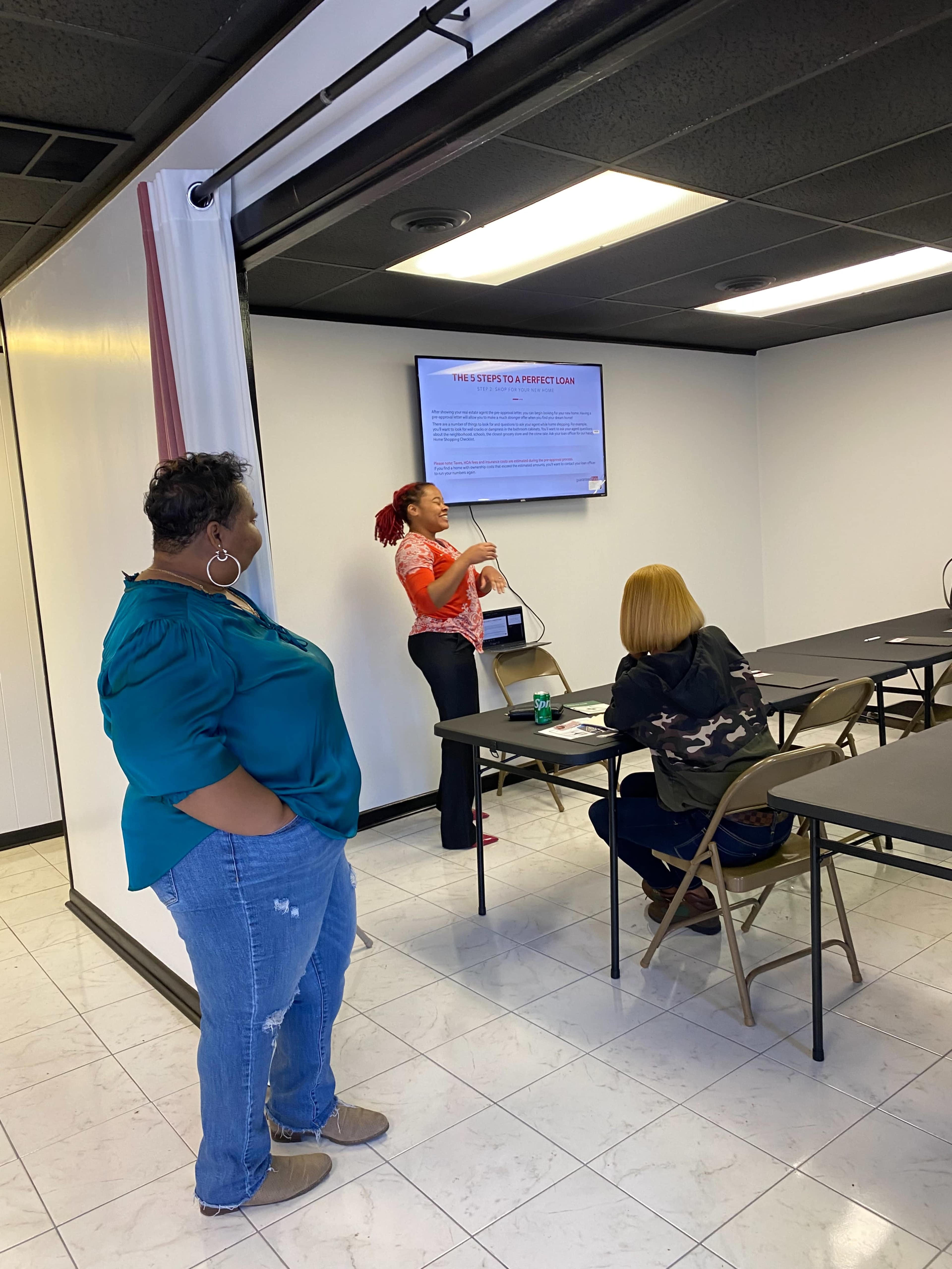 A woman stands at the front of a classroom, presenting information on a screen while two seated individuals listen attentively.