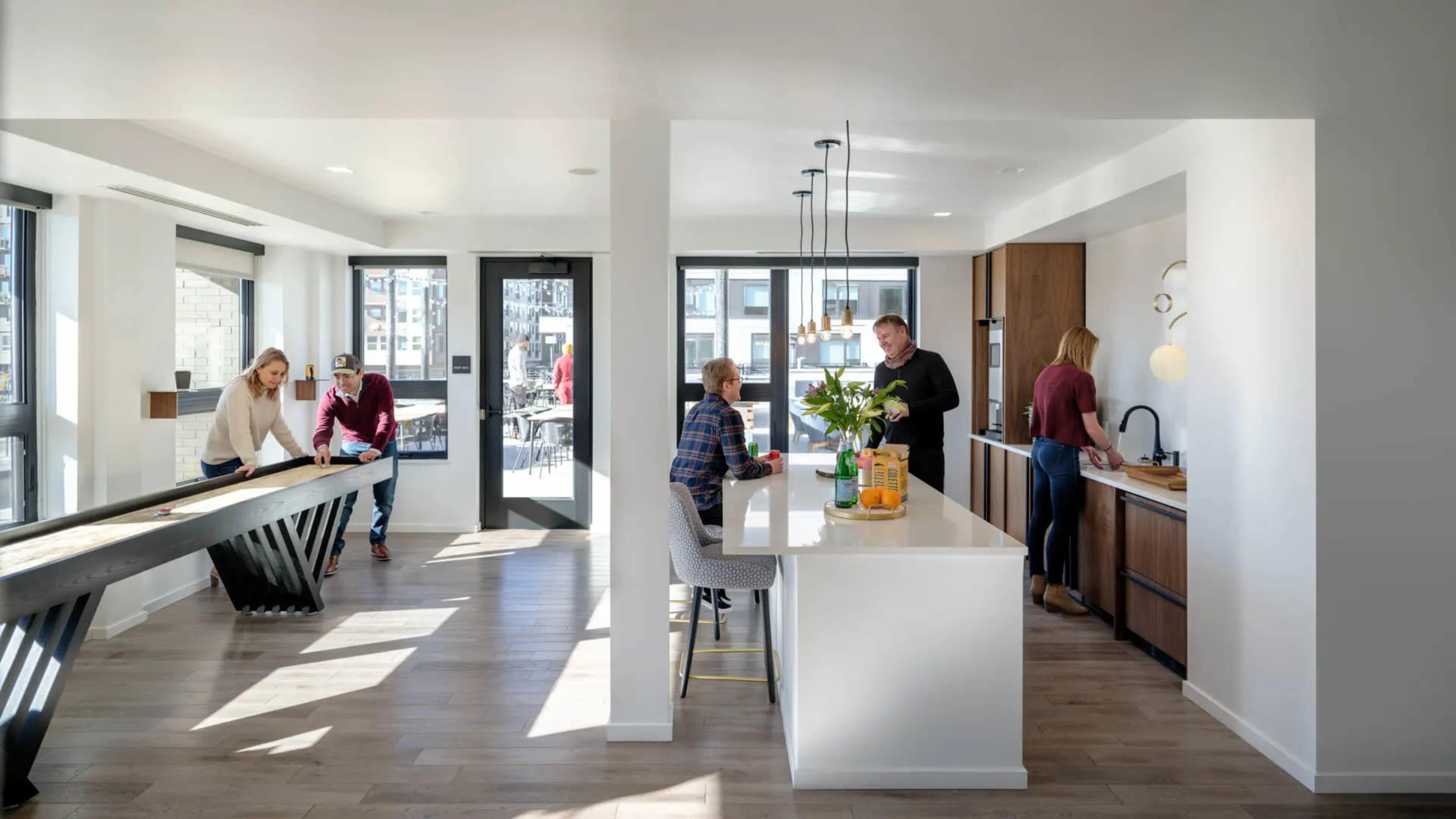 A modern open-concept kitchen and dining area features a large island, a dining table, and two people preparing food while others socialize nearby.