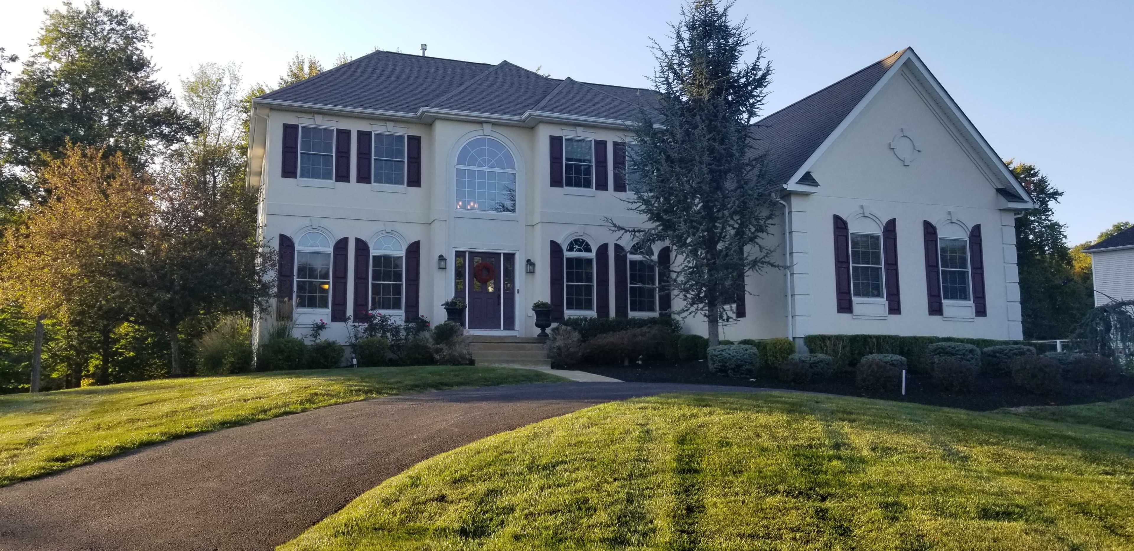 A large two-story house with a symmetrical facade, multiple windows, and a sloping green lawn is situated on a paved driveway.