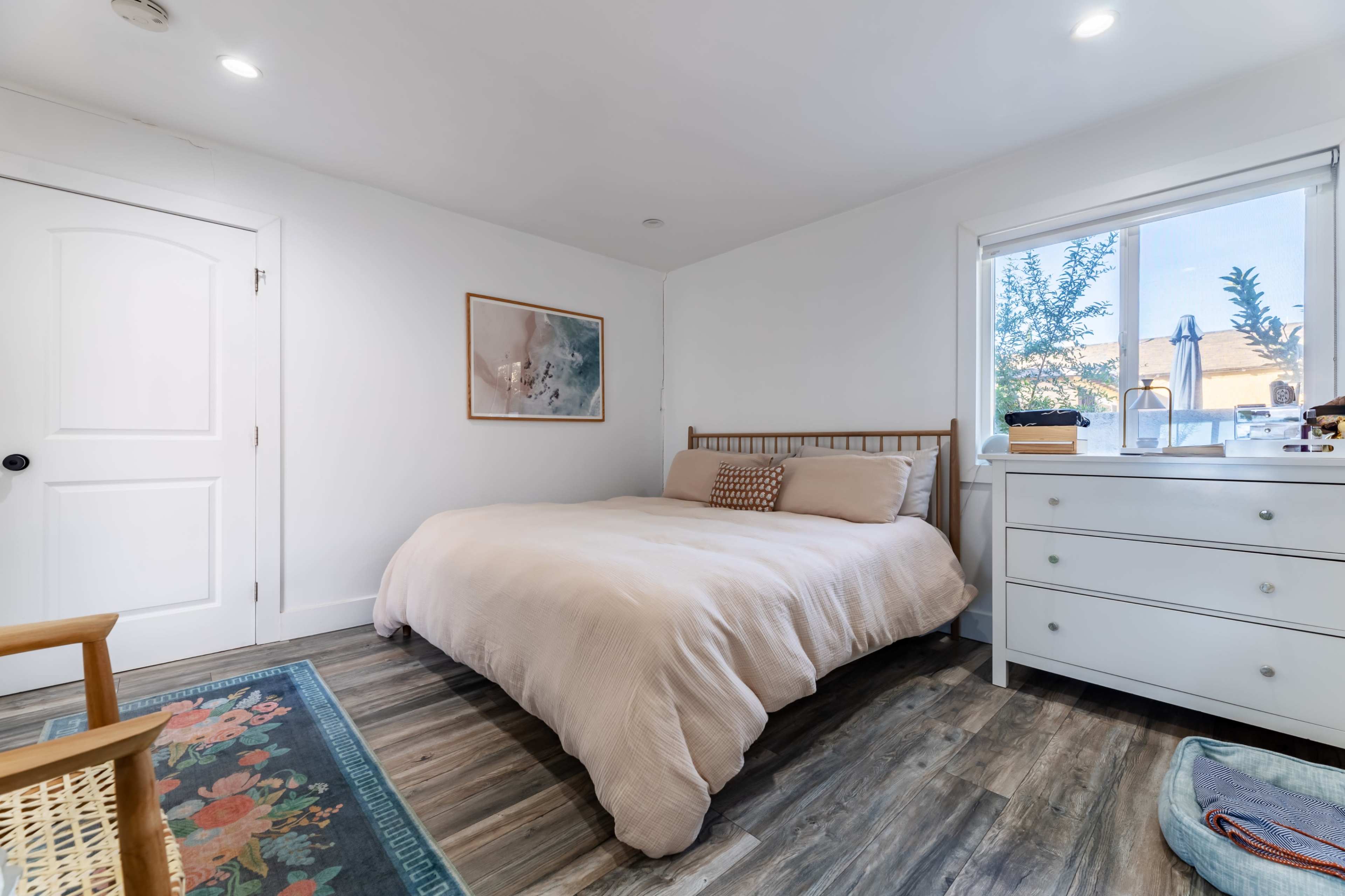 The image shows a neatly arranged bedroom featuring a bed with a light-colored cover, a window with natural light, and a dresser next to it.