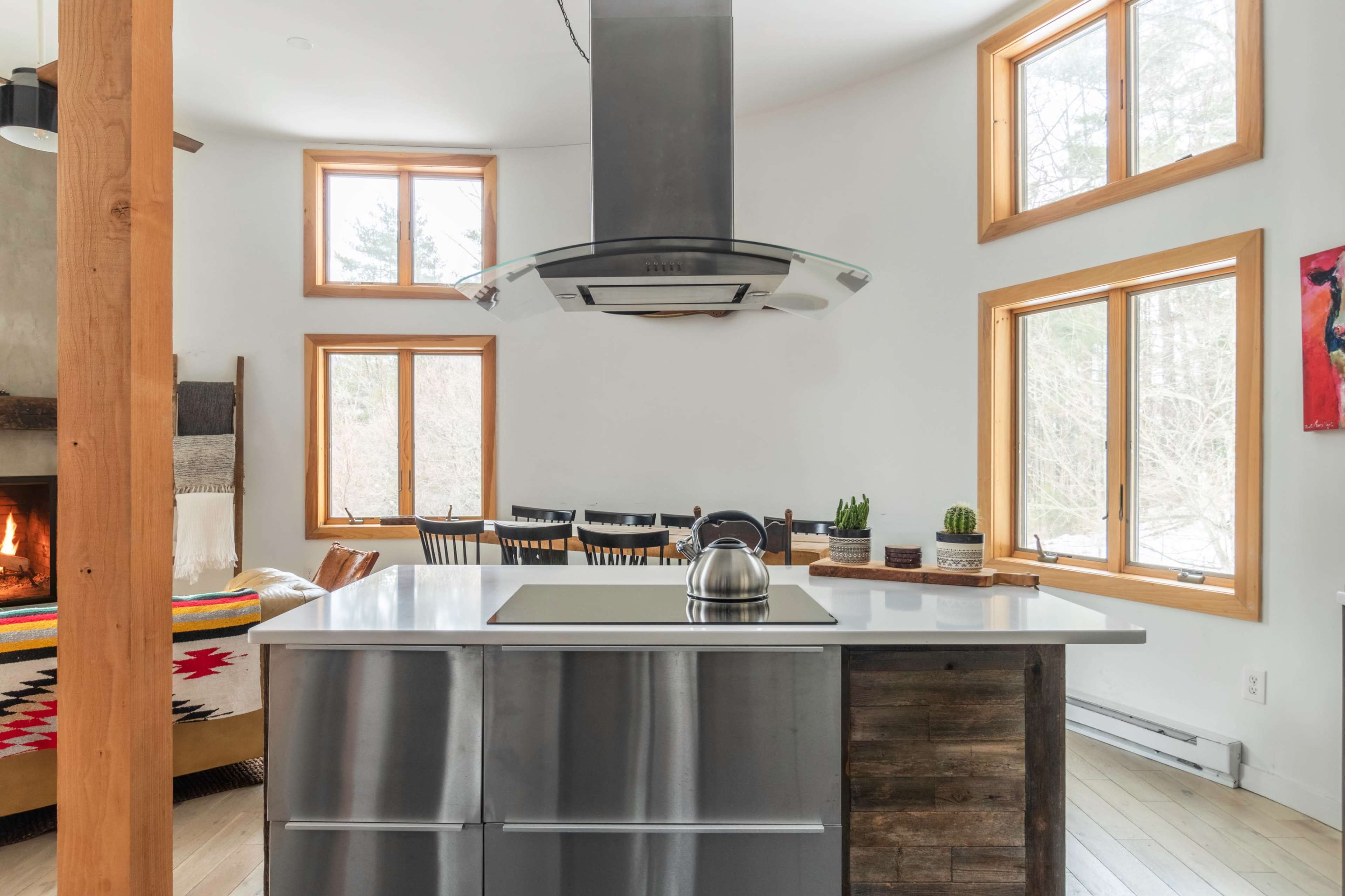 A modern kitchen features a central stovetop island with a hood, large windows, and a cozy seating area with a fireplace.