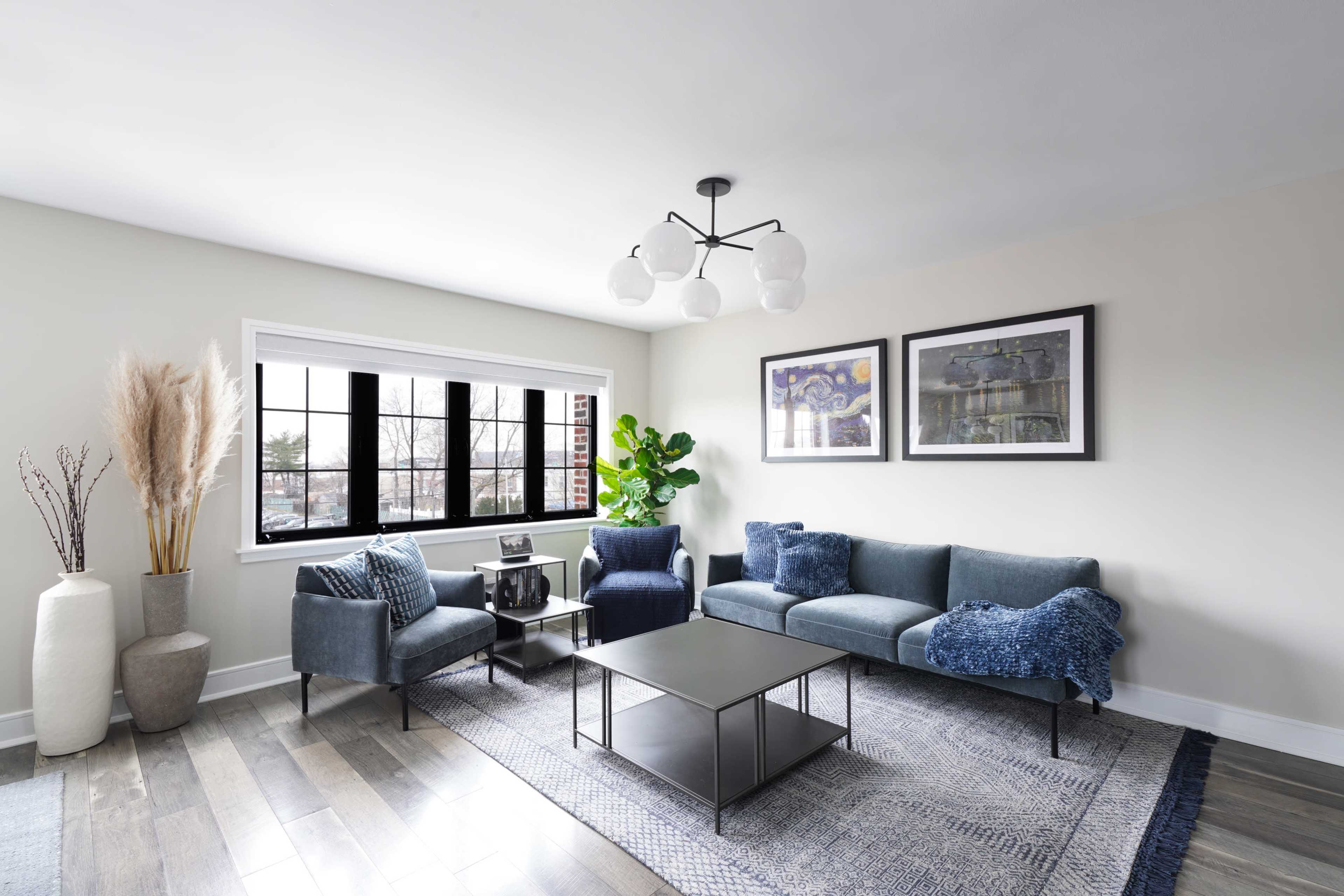 A modern living room featuring a gray sofa, a black coffee table, artwork on the walls, and a large window with indoor plants.