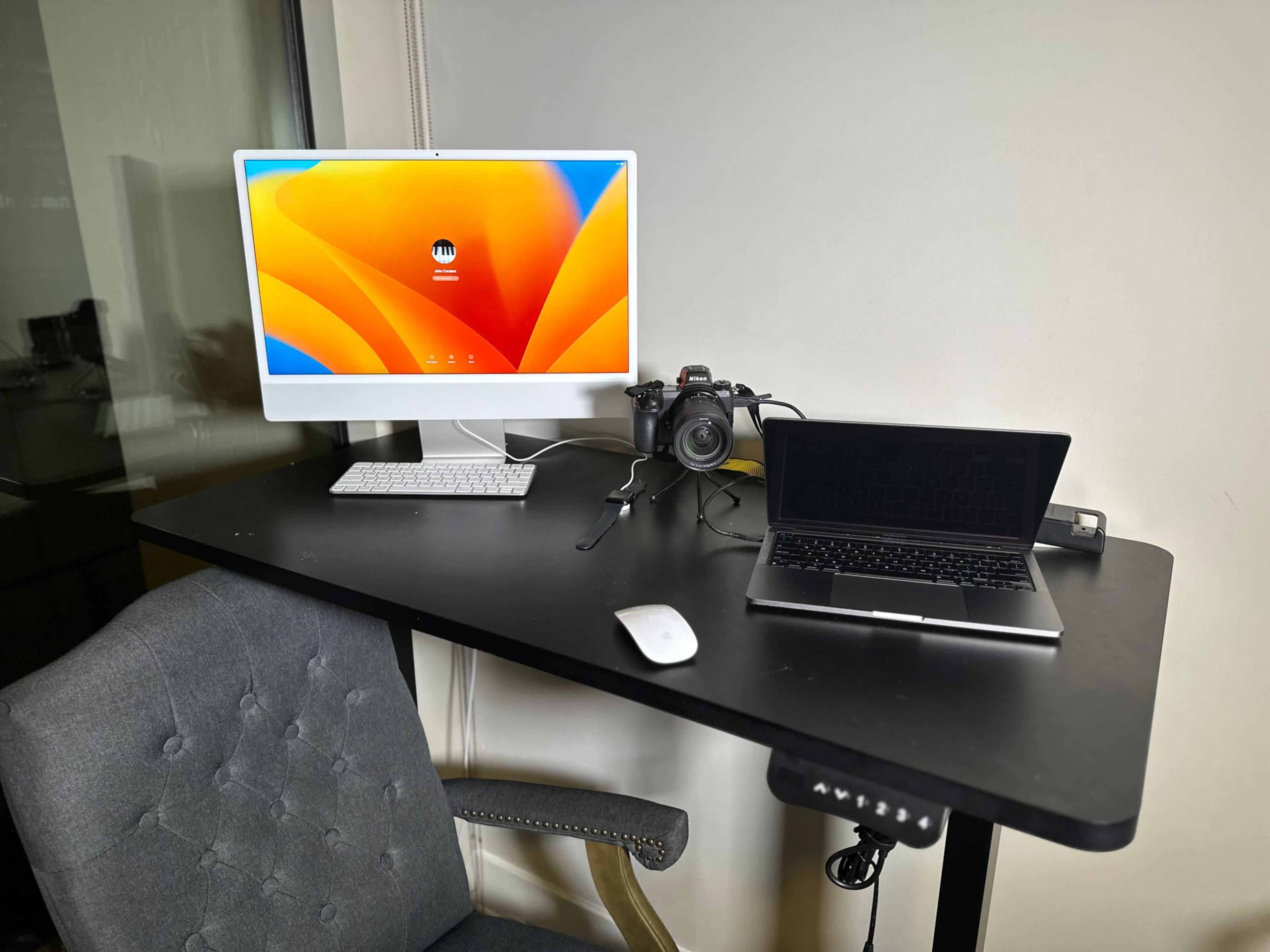 A desktop setup featuring a large monitor, a laptop, a camera, and a wireless keyboard and mouse on a black table with a gray chair.