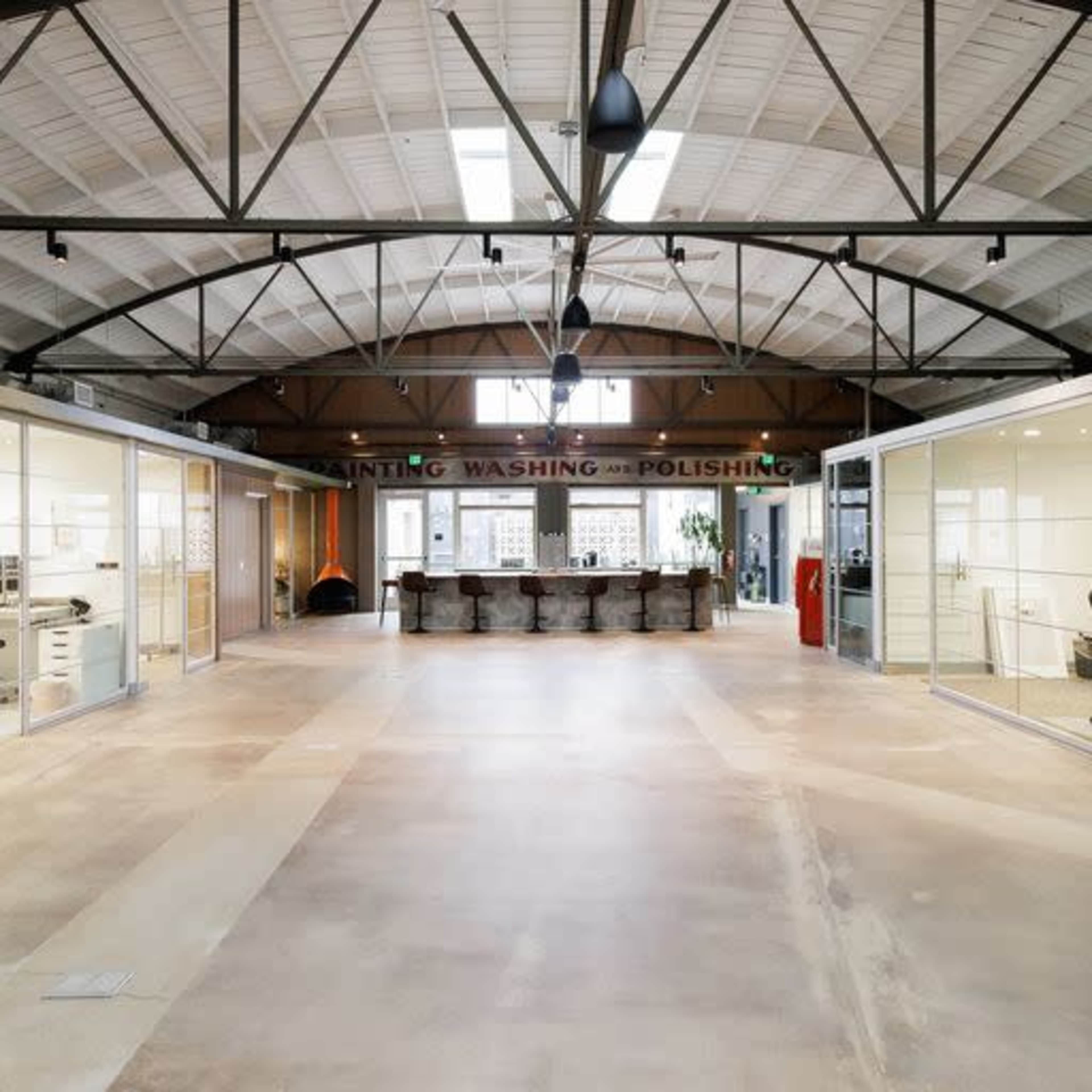 A spacious industrial-style office with high ceilings, large glass cubicles on either side, and a central area featuring a communal table.