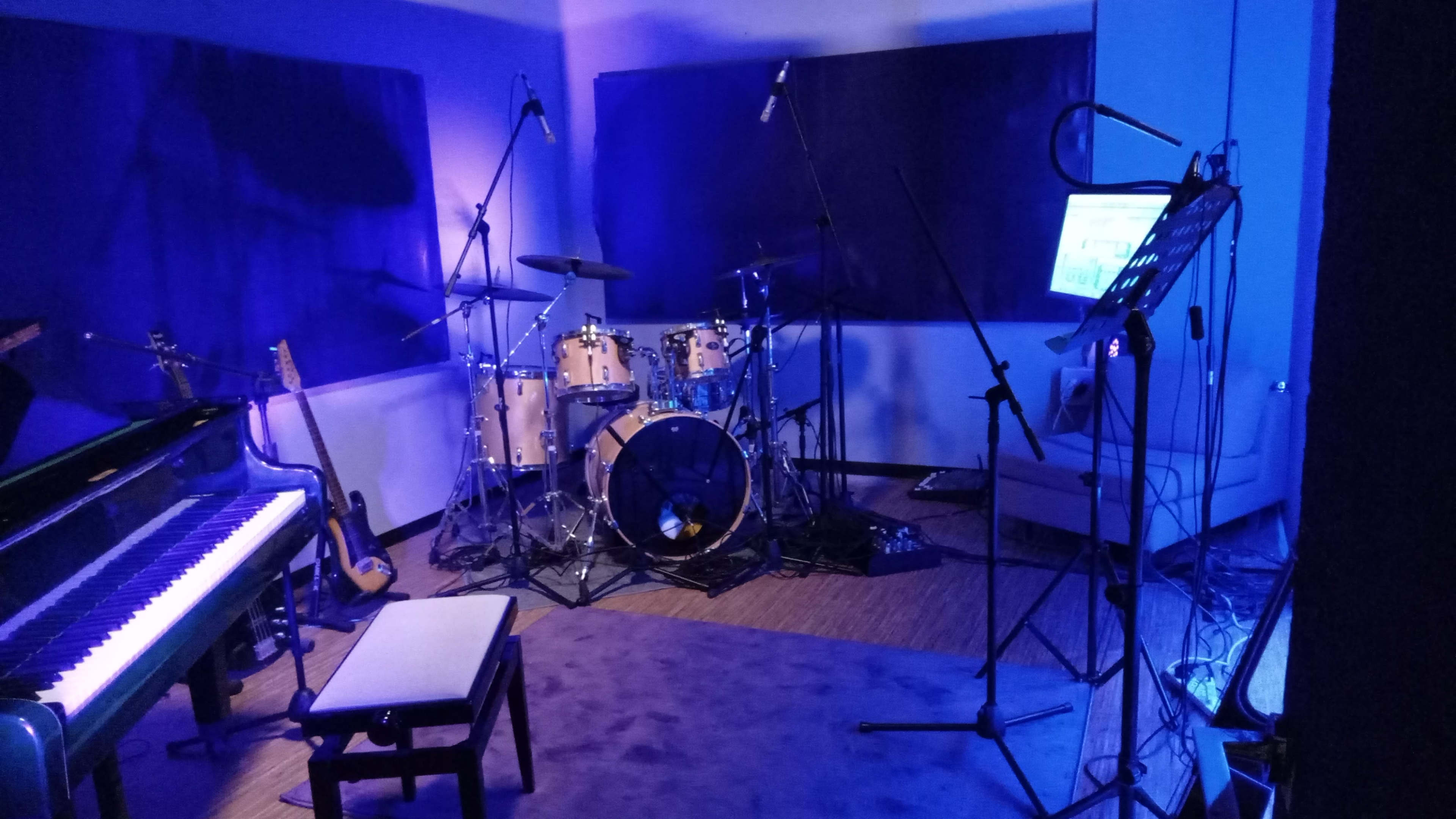 A music studio setup with a piano, drum kit, and microphone stands under blue lighting.