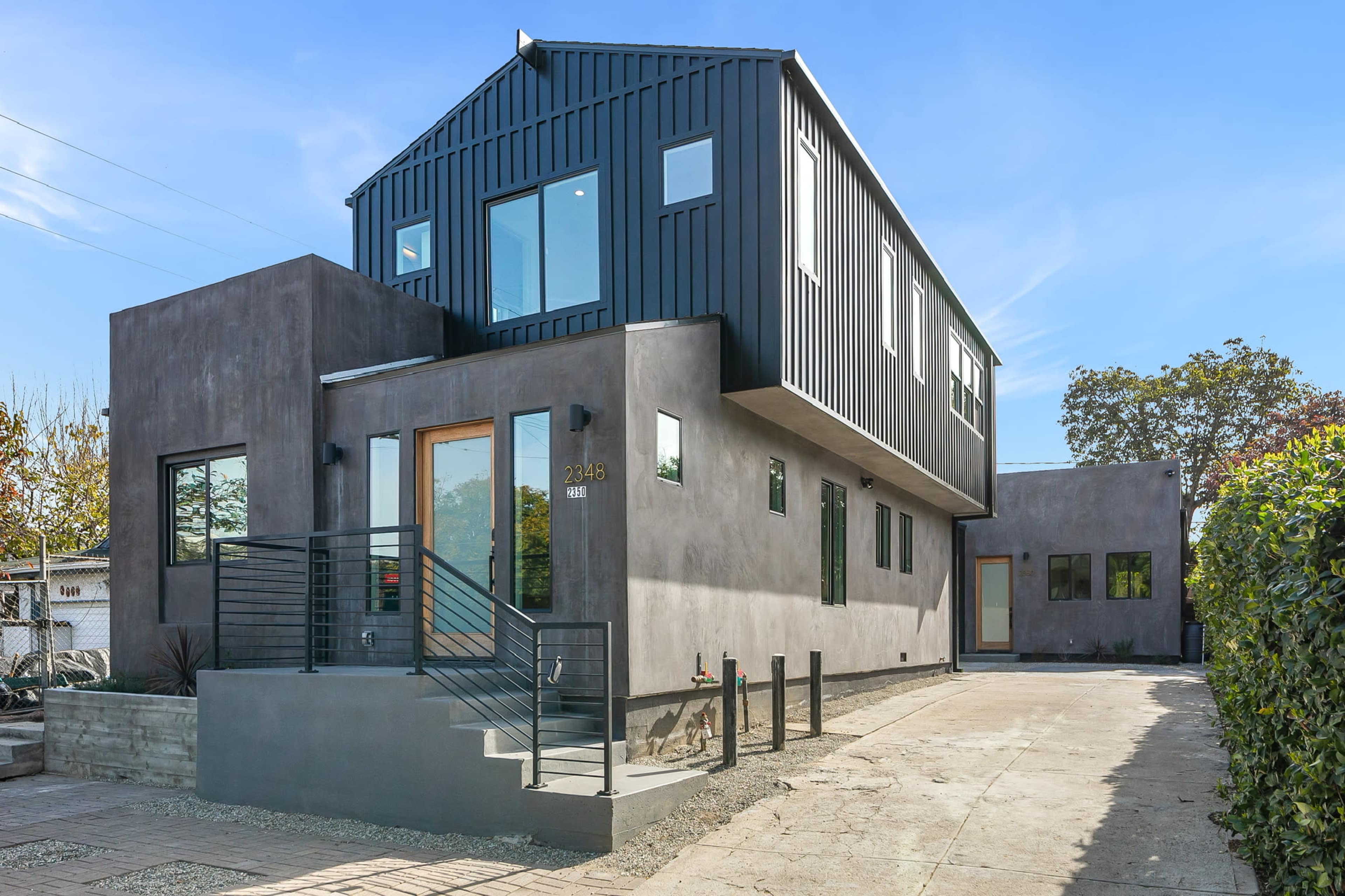 A modern two-story house with a dark metal facade and a concrete exterior, featuring large windows and a landscaped entrance.