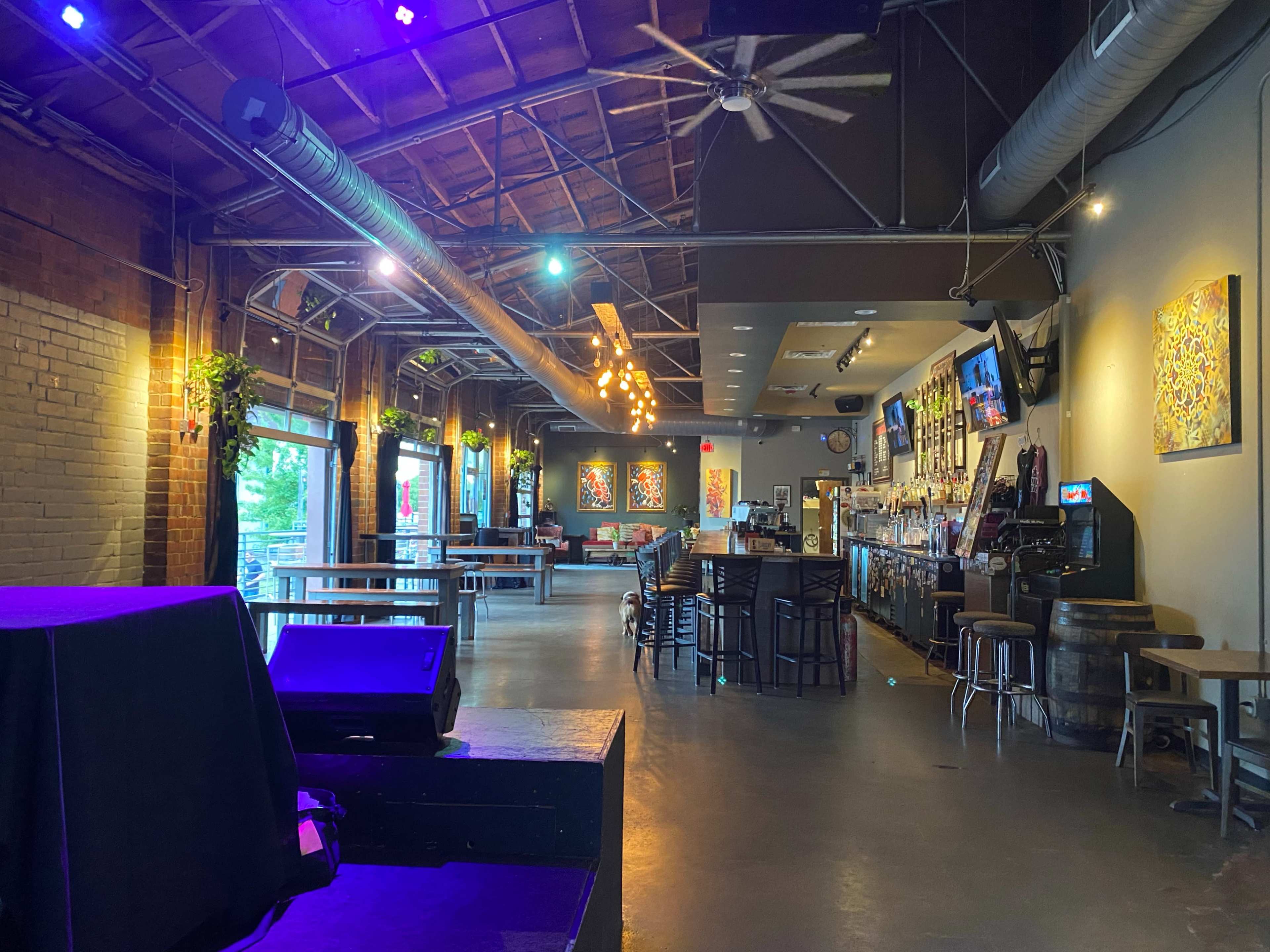 The interior of a spacious bar with wooden accents, a long bar counter, seating arrangements, and colorful artwork on the walls.