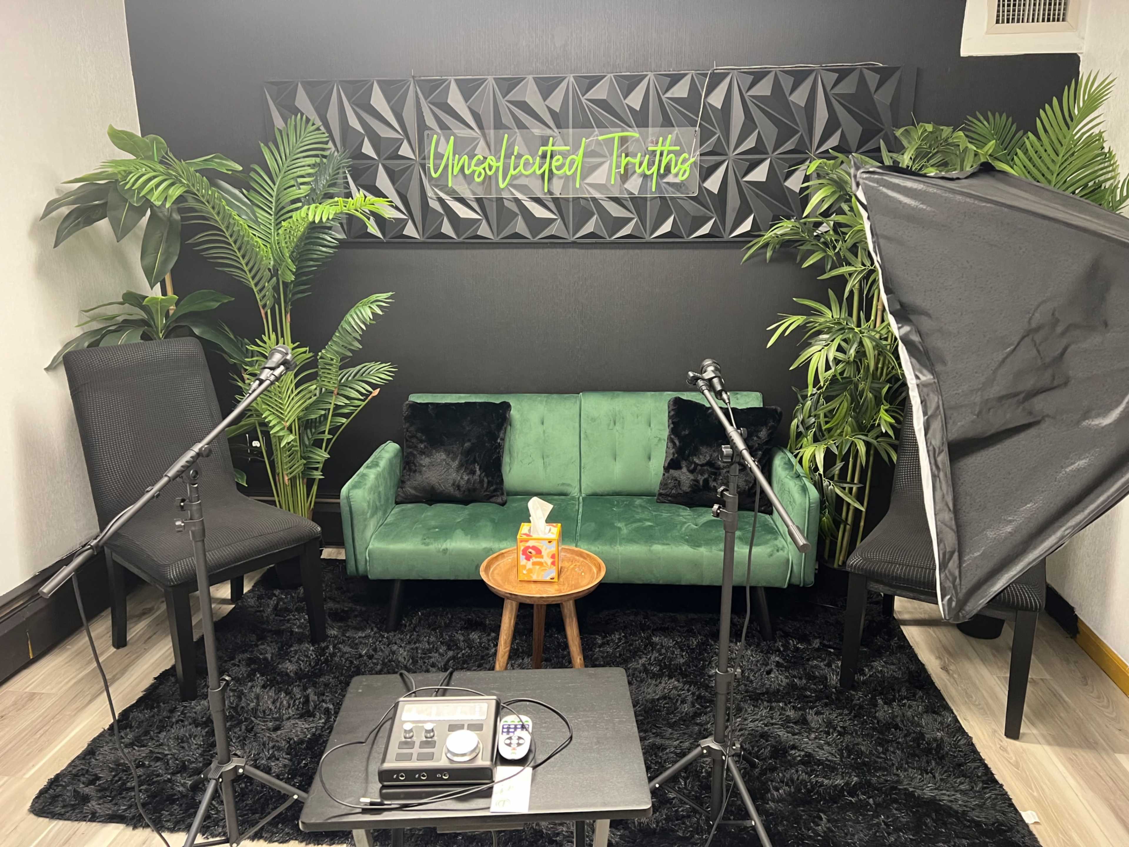 A modern studio setup with a green sofa, a small wooden table, two lighting setups, and decorative plants against a wall featuring a sign that reads "Unsolicited Truths."