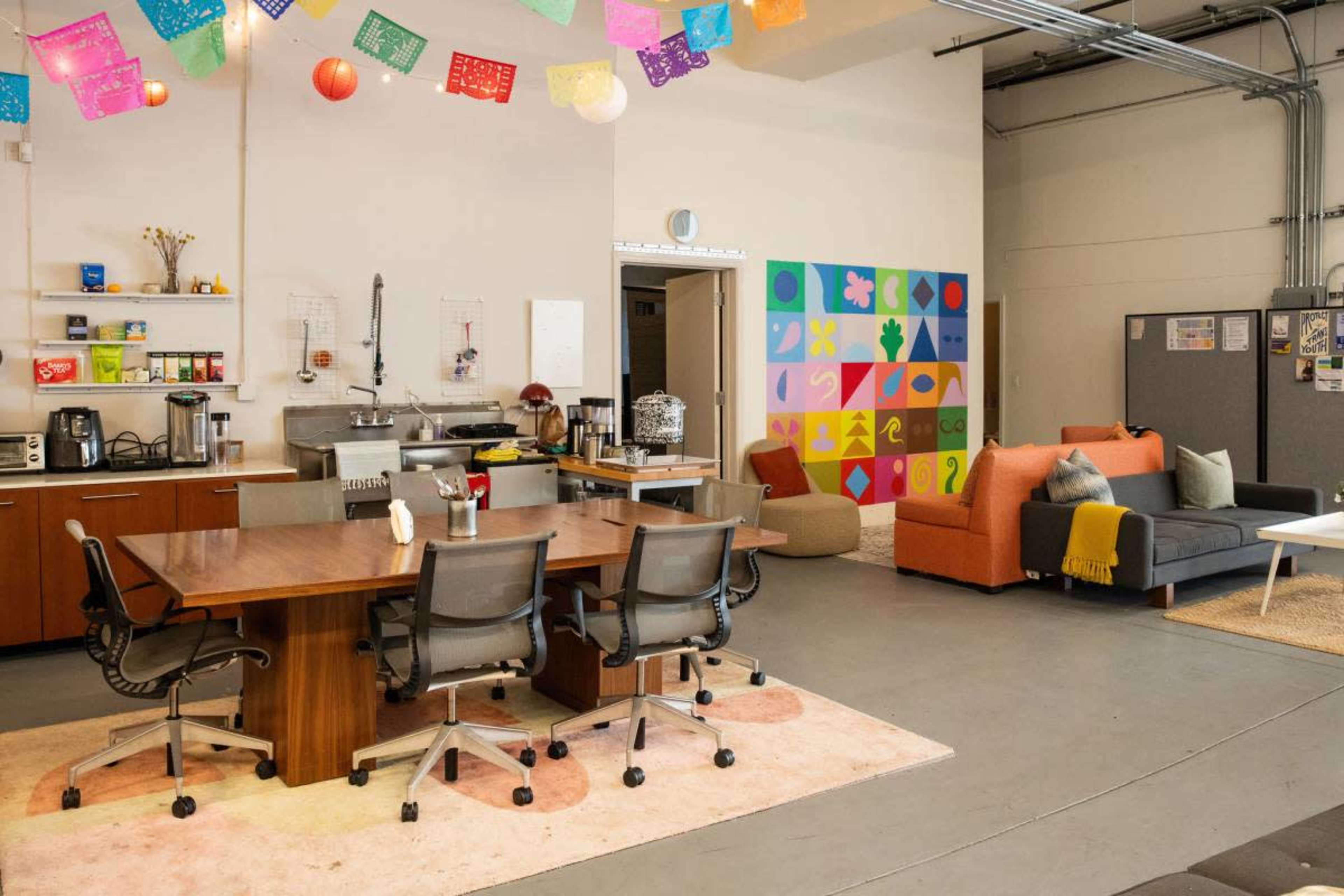 The image shows a modern communal space with a kitchen area featuring a large table and chairs, and a lounge area with colorful couches and wall decorations.