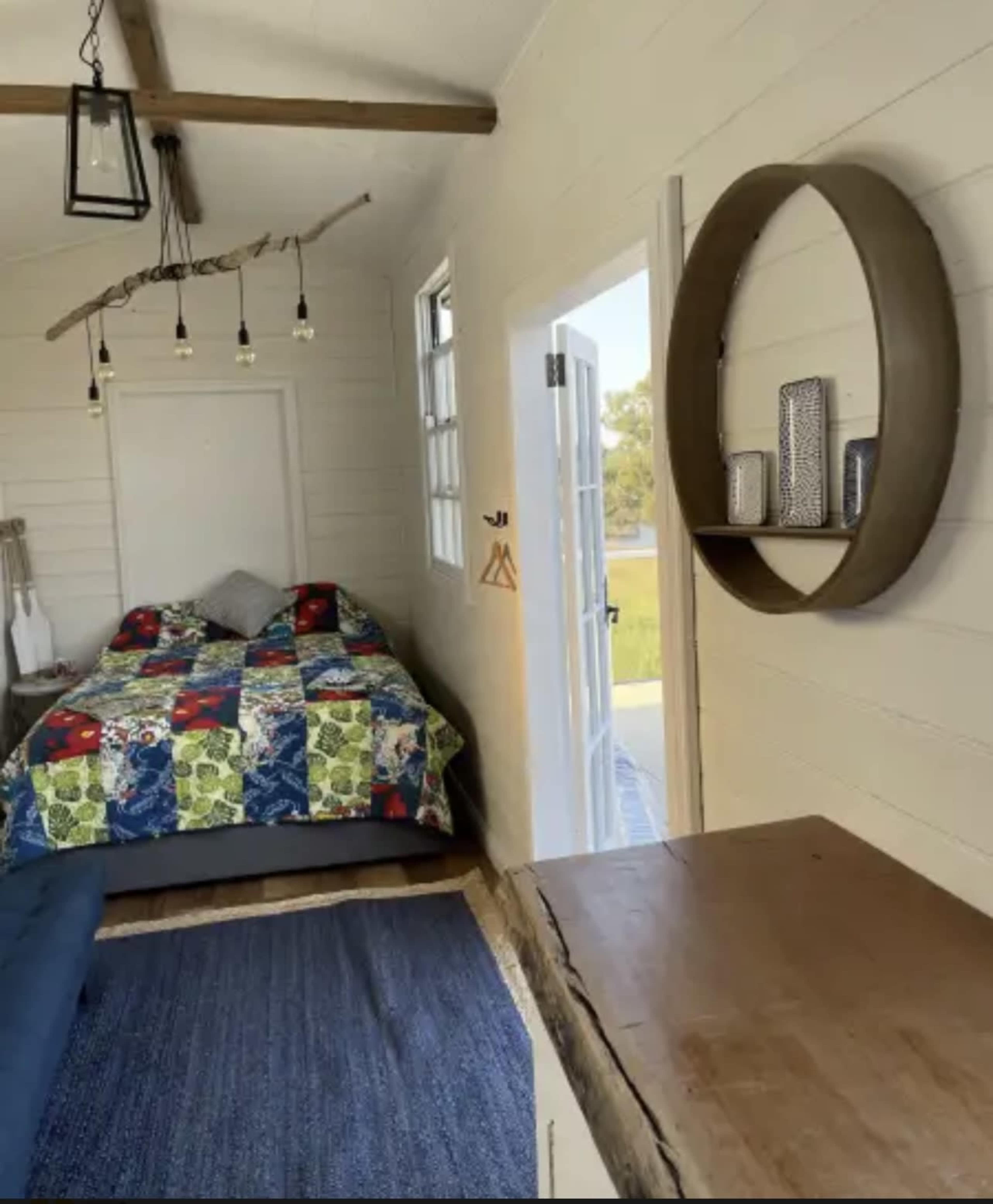 A cozy bedroom with a patterned quilt on the bed, a small seating area, and decorative items on a circular wall shelf.