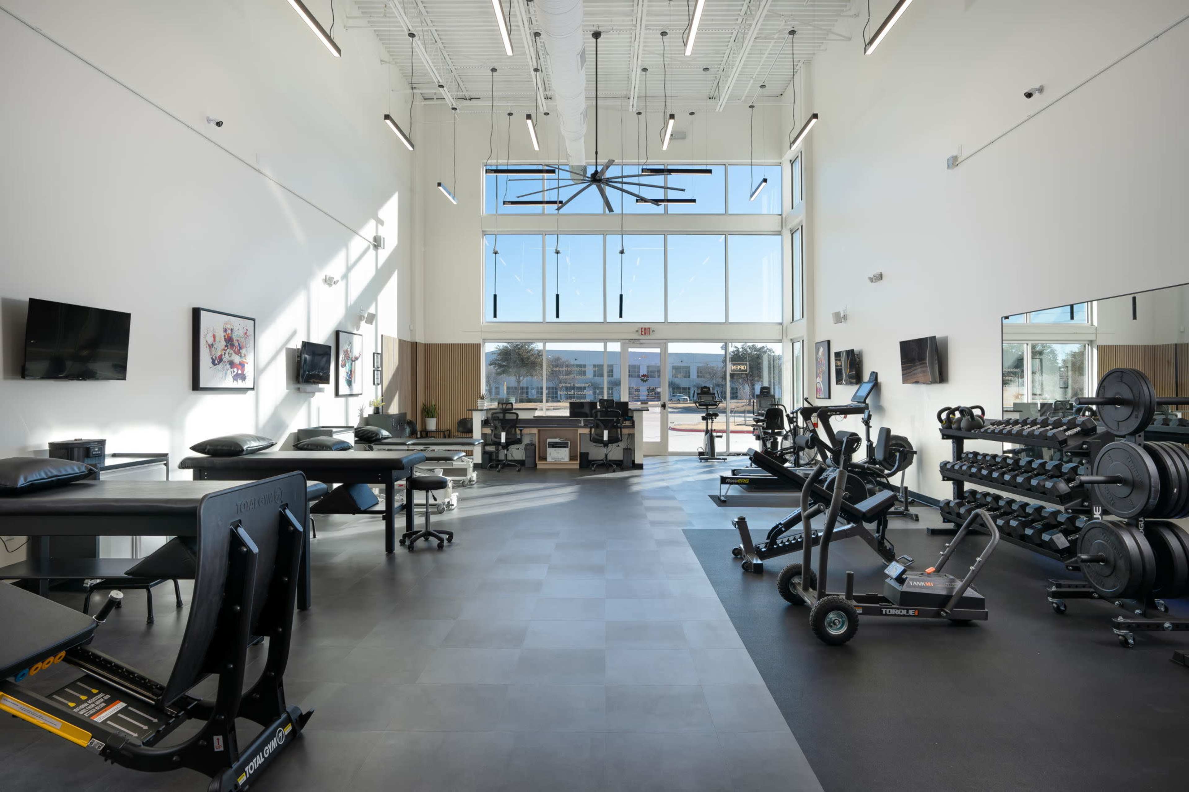 A modern fitness facility with exercise equipment, including weight racks and a cardio machine, under large windows that provide ample natural light.