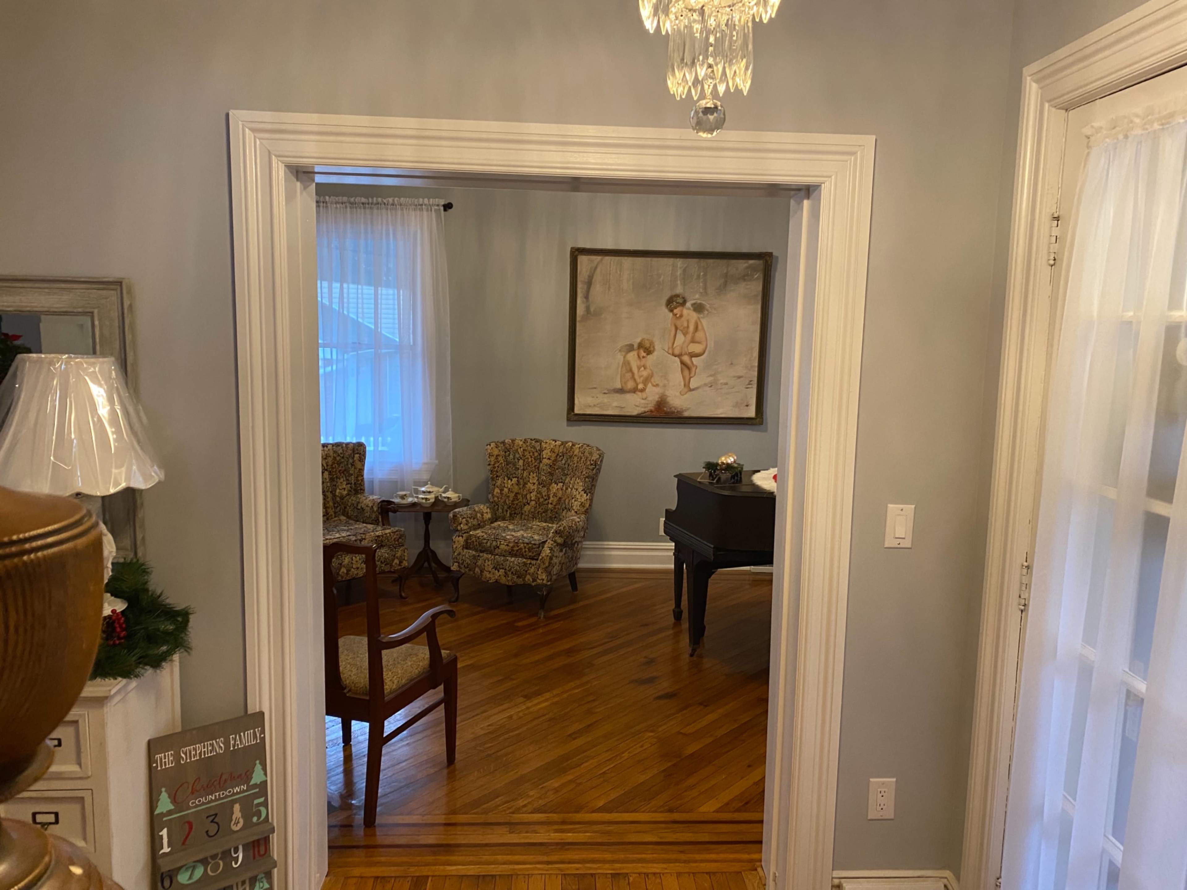 A view through a doorway shows a cozy sitting area with two patterned chairs and a piano, complemented by a large painting on the wall.