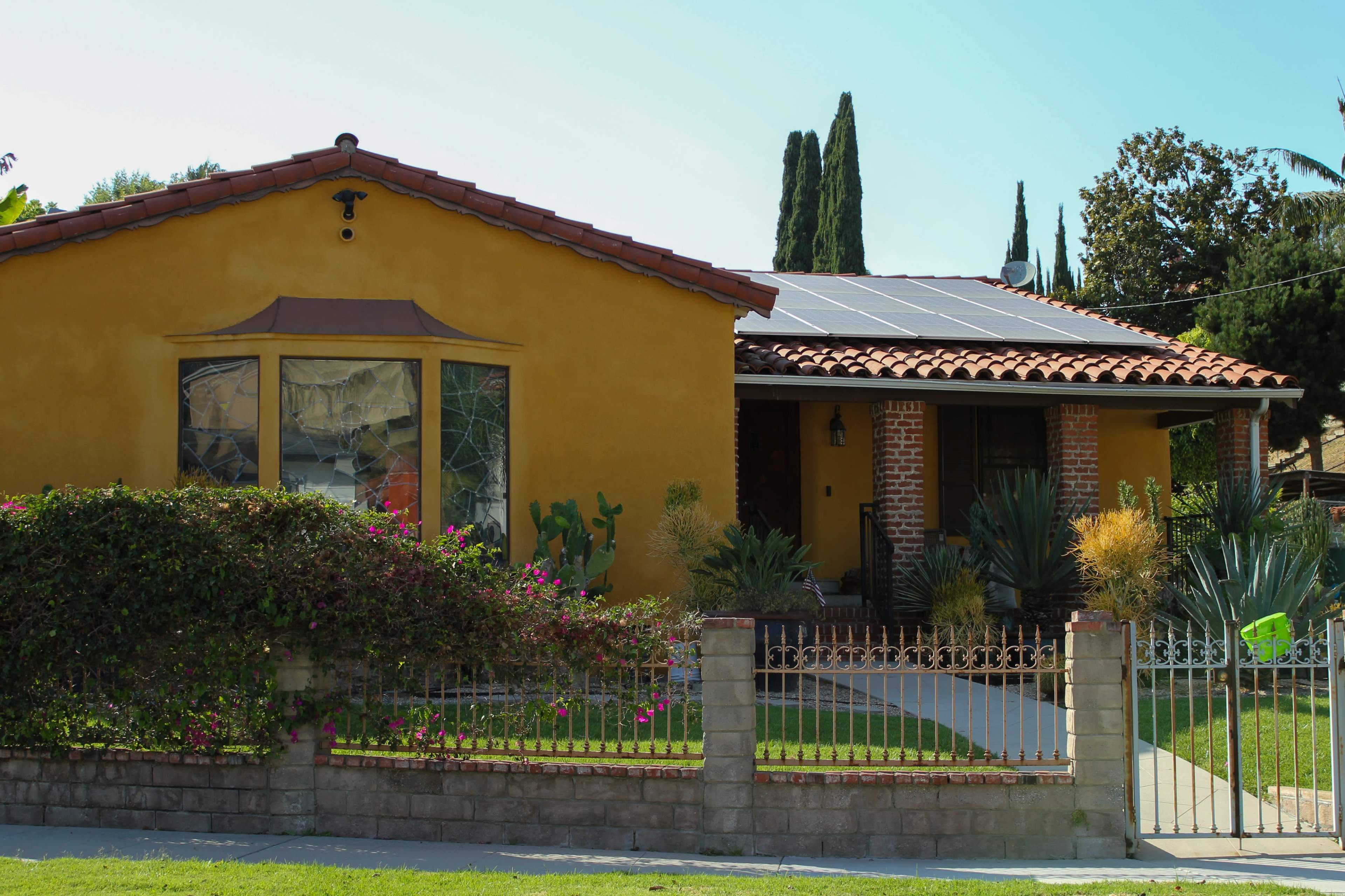 A single-story house with a terracotta tile roof features solar panels, surrounded by a garden and a decorative fence.