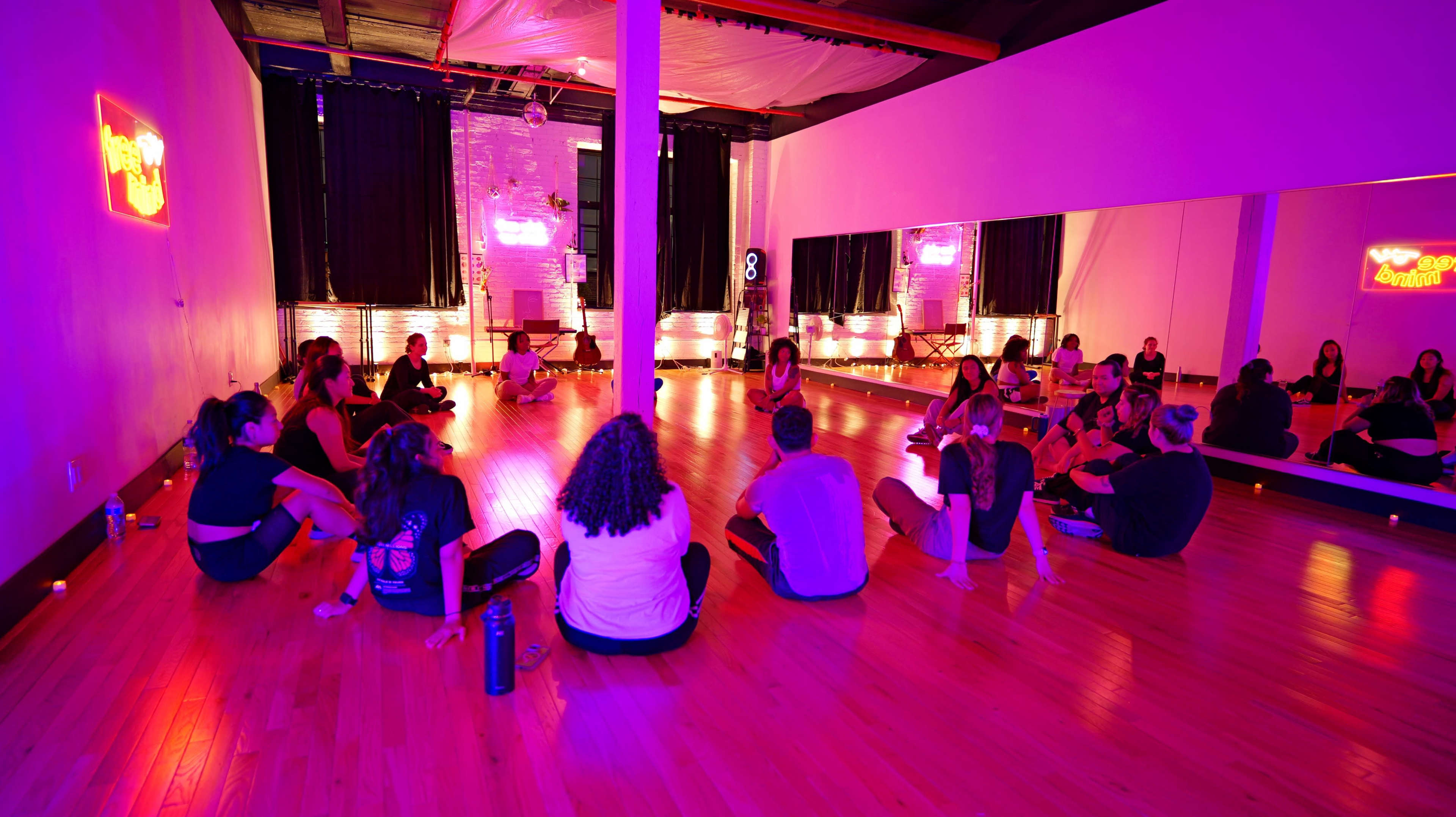 A group of people sits in a circle on a wooden floor in a brightly lit yoga studio with soft lighting and mirrors.
