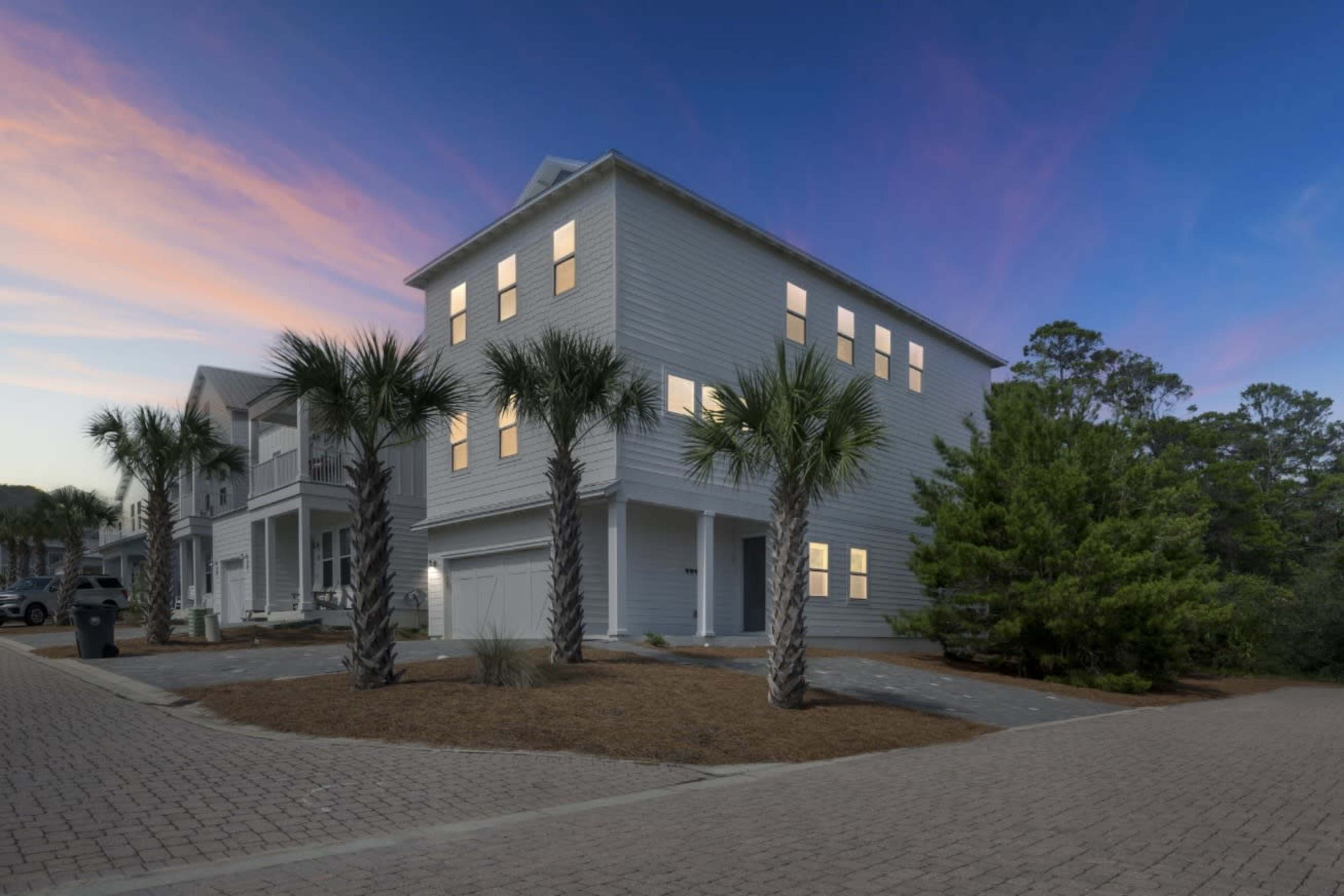 A modern, two-story coastal home with large windows is surrounded by palm trees and set against a colorful sunset.