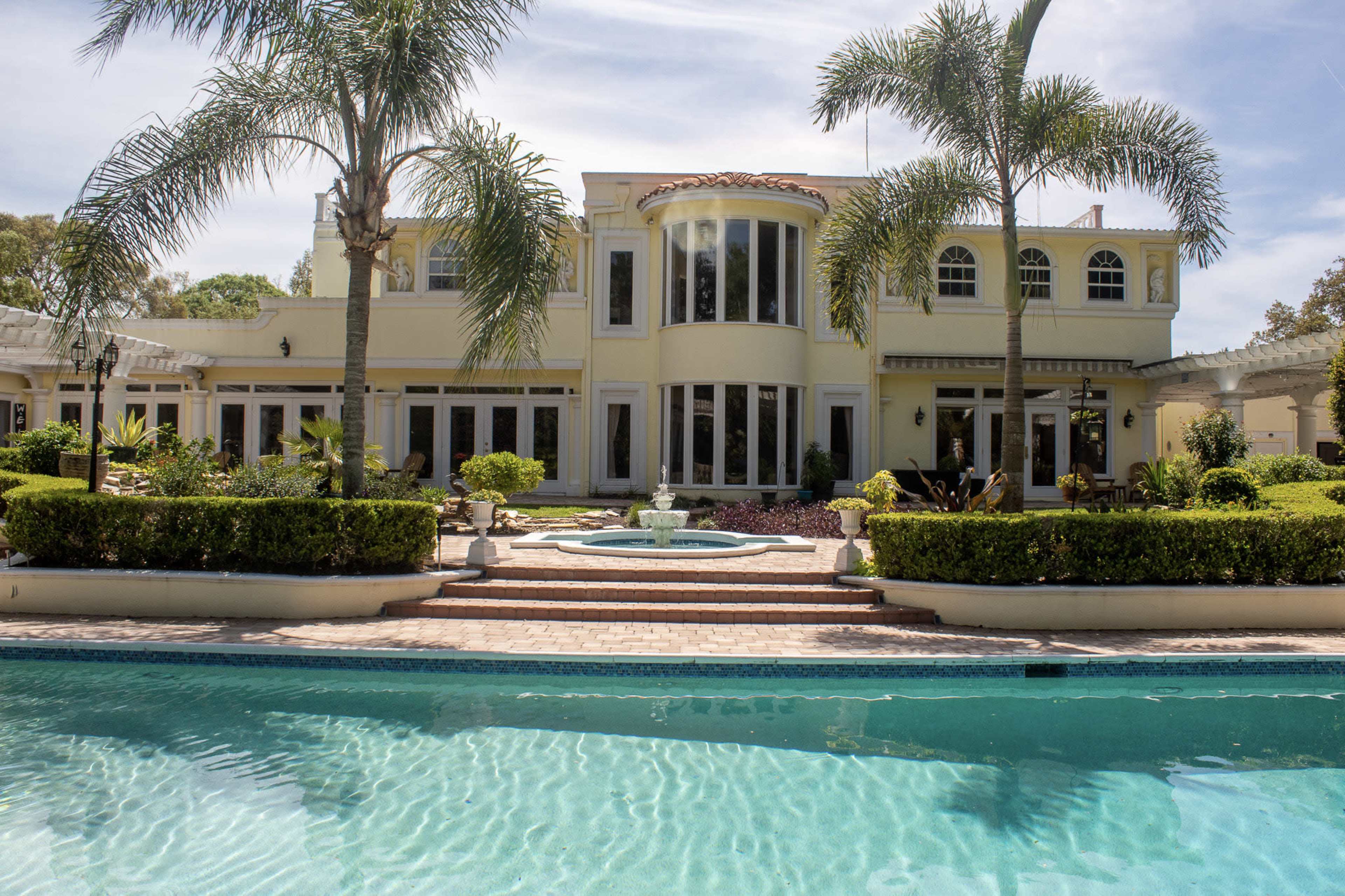 A large, modern house with multiple windows and a pool in the foreground, surrounded by neatly maintained landscaping and palm trees.