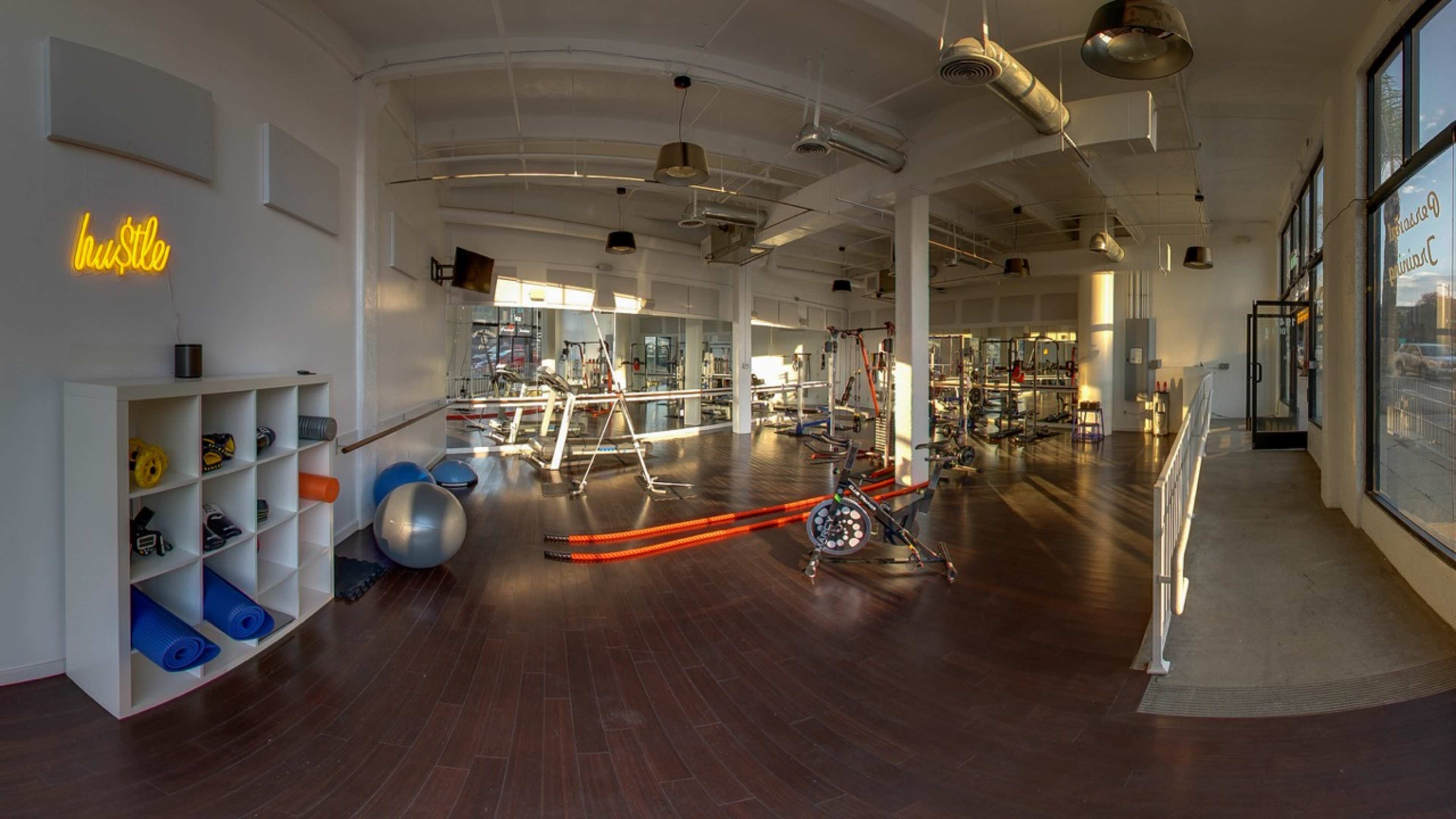 A spacious gym interior featuring exercise equipment, mats, and a wooden floor, with natural light coming through large windows.