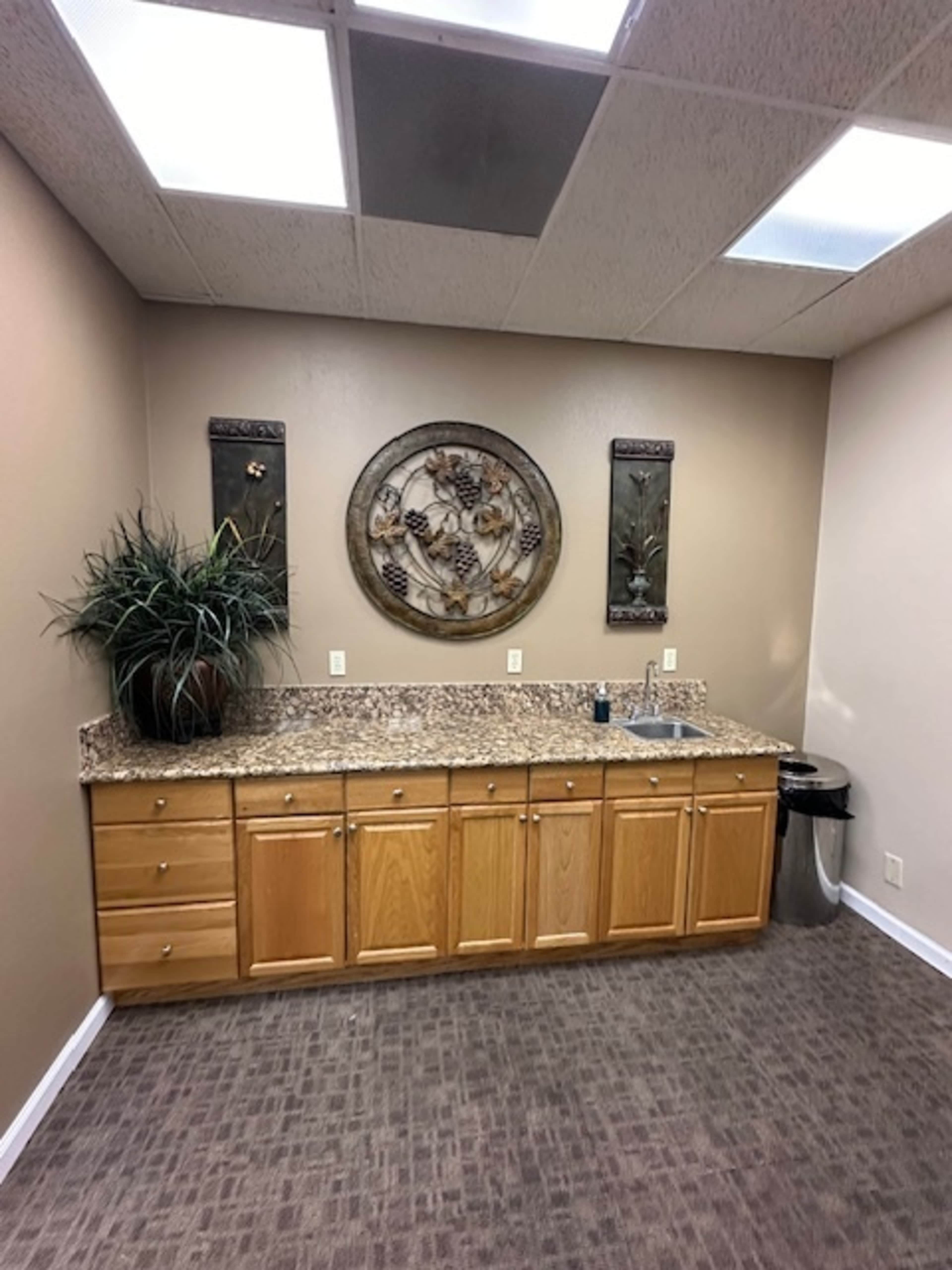 A small kitchenette with wooden cabinets, a granite countertop, a sink, and decorative wall art, set against a neutral-toned wall.