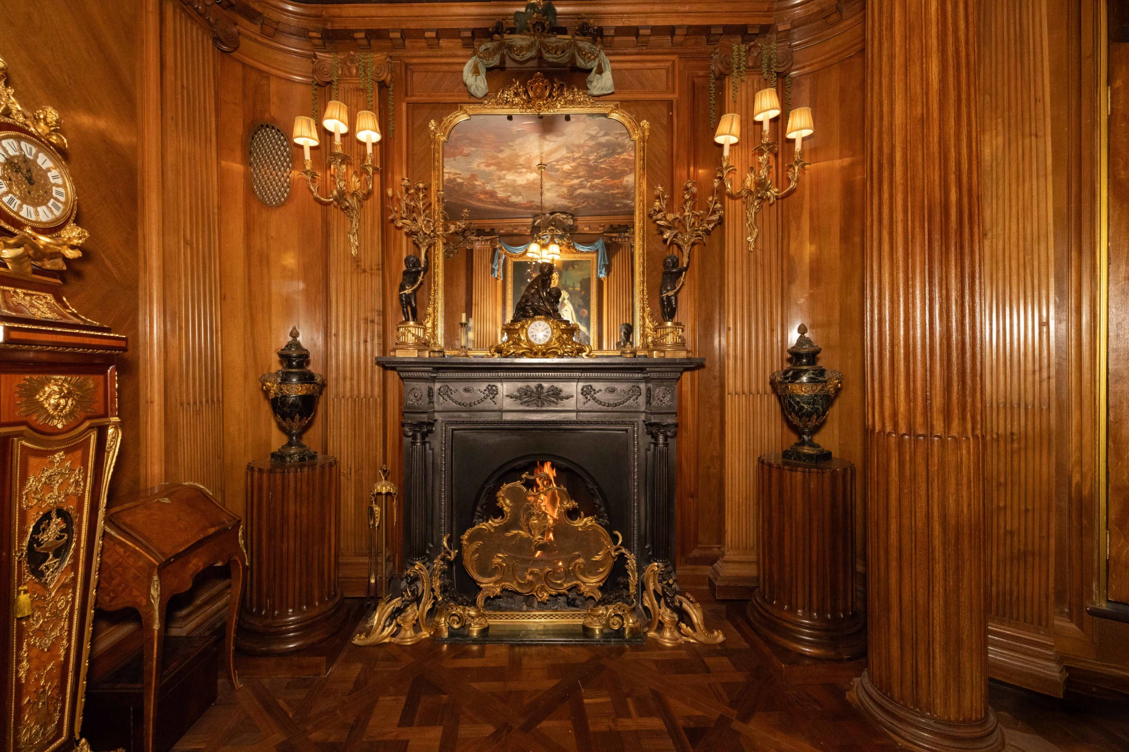 The image features an ornate fireplace surrounded by wooden paneling, mirrors, and decorative wall sconces in a richly detailed room.