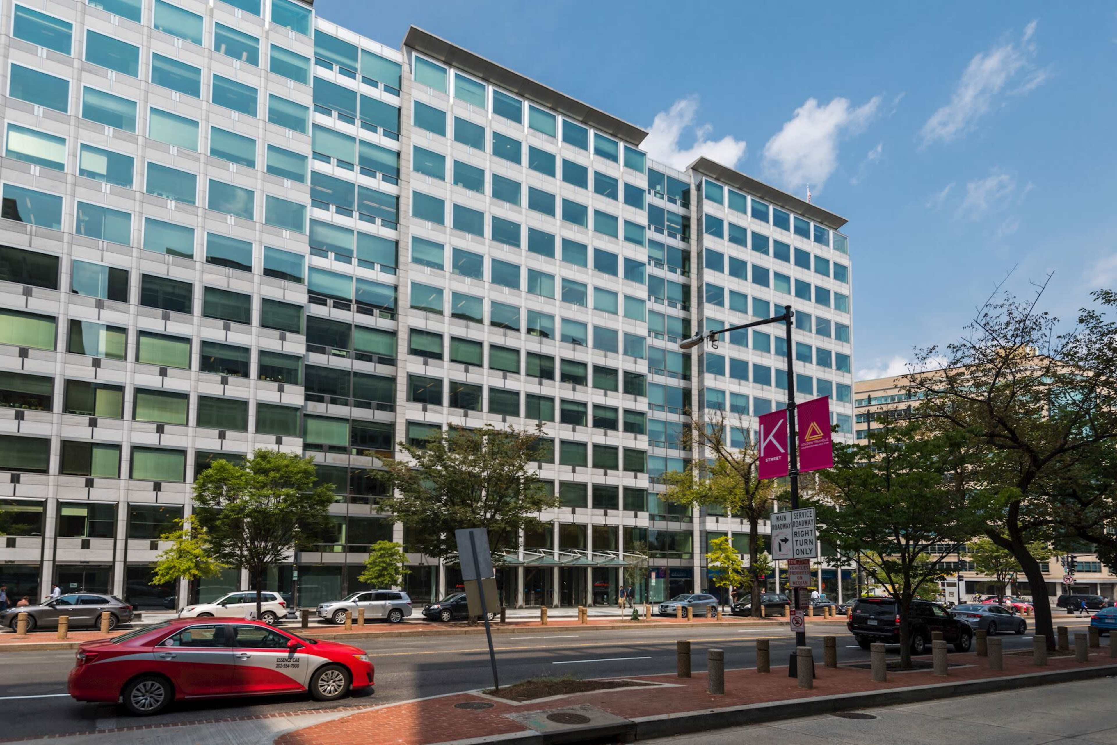 A modern glass-faced office building is situated on a busy street lined with trees and parked cars.
