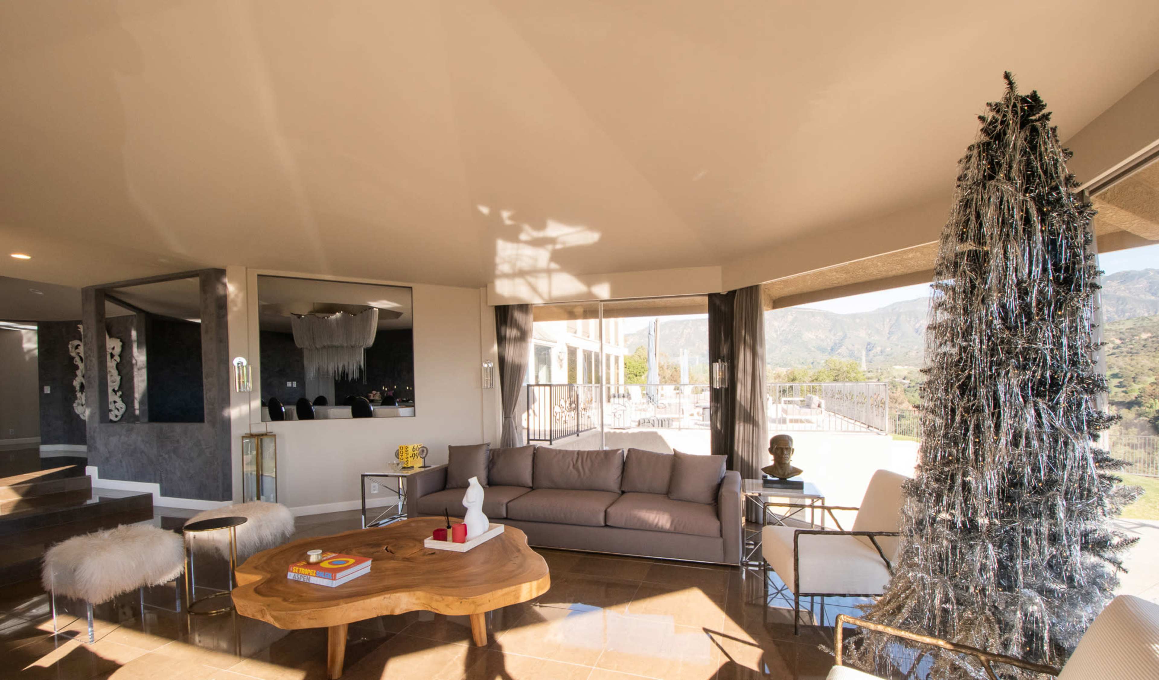 The living room features a modern design with a wooden coffee table, a gray sofa, and a decorated Christmas tree, all set against a backdrop of large windows showing a mountainous view.
