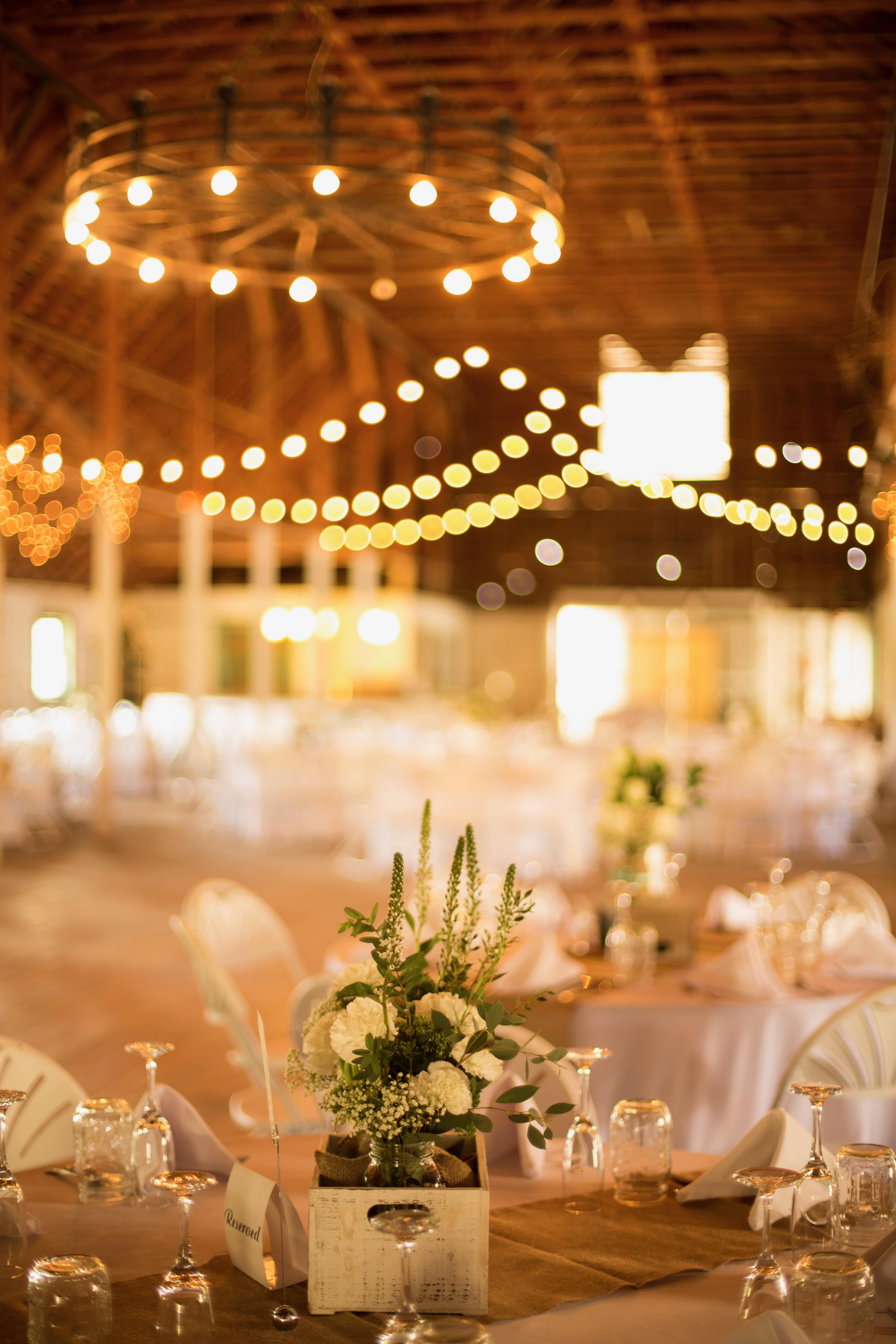 A rustic event space features neatly arranged tables with white linens, a centerpiece of flowers in a wooden box, and string lights illuminating the ceiling.