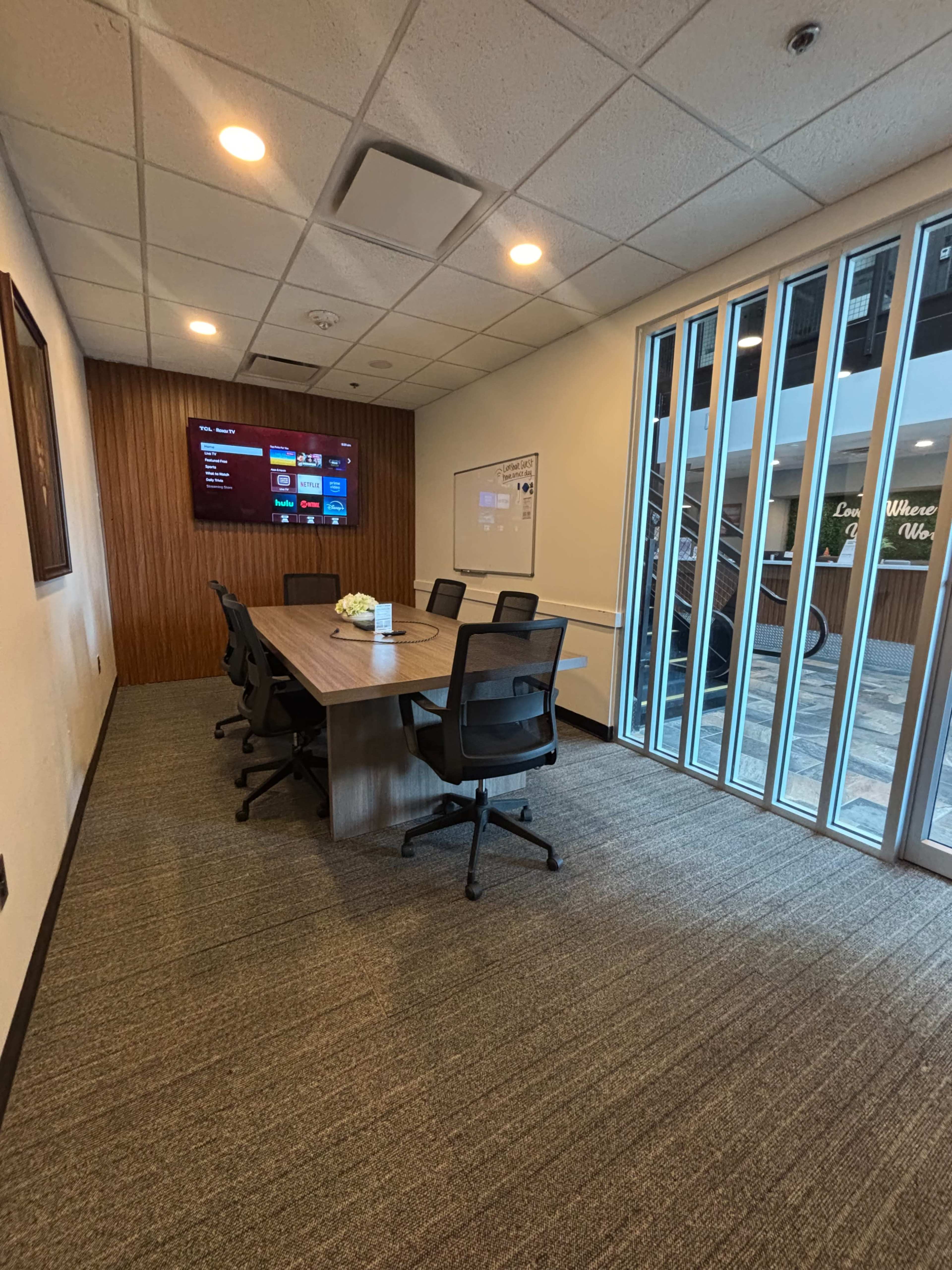 The image shows a conference room with a long table and six chairs, a wall-mounted screen displaying information, and large windows overlooking an outdoor area.