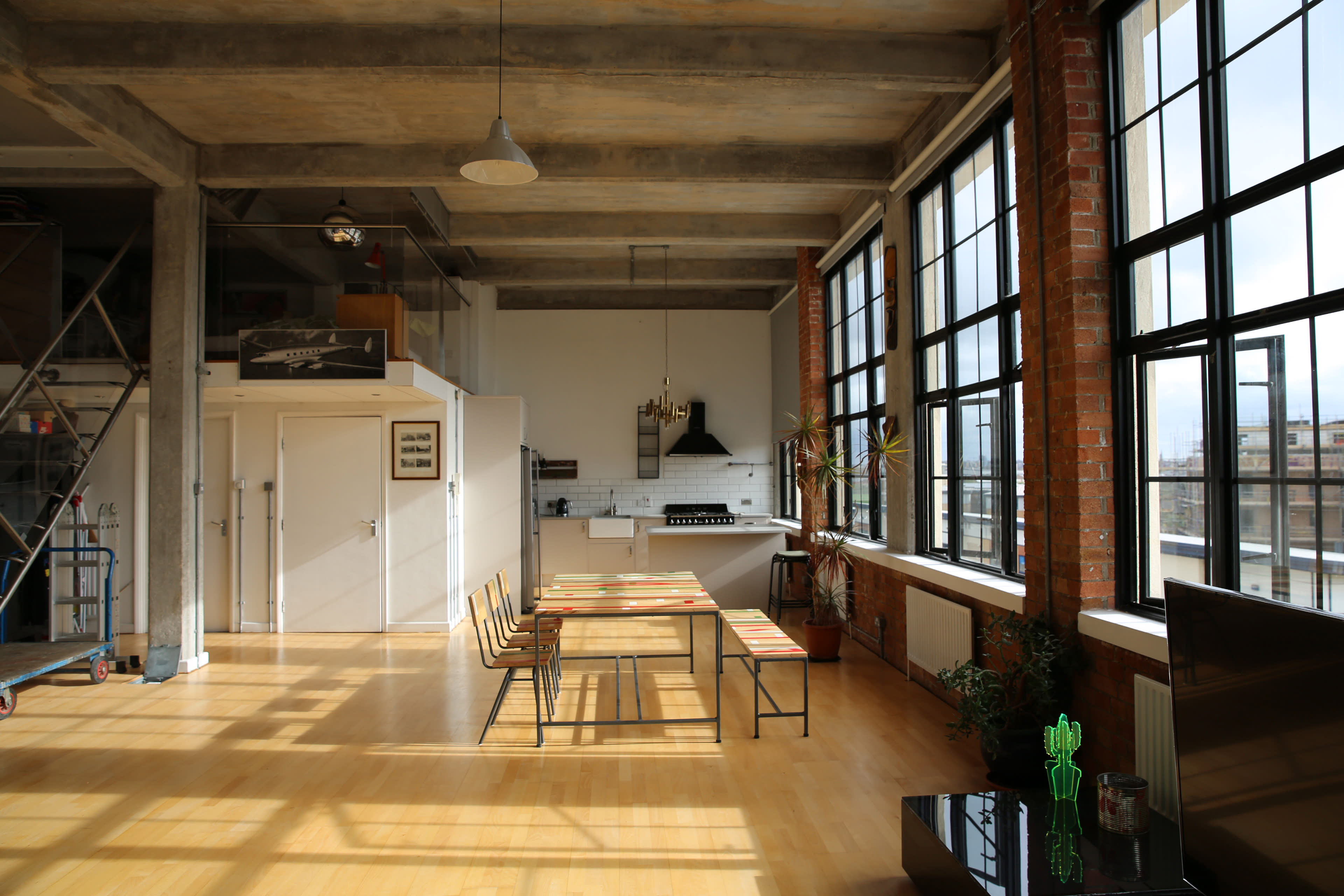 A spacious loft features large windows, a wooden dining table with chairs, and a kitchen area in the background.