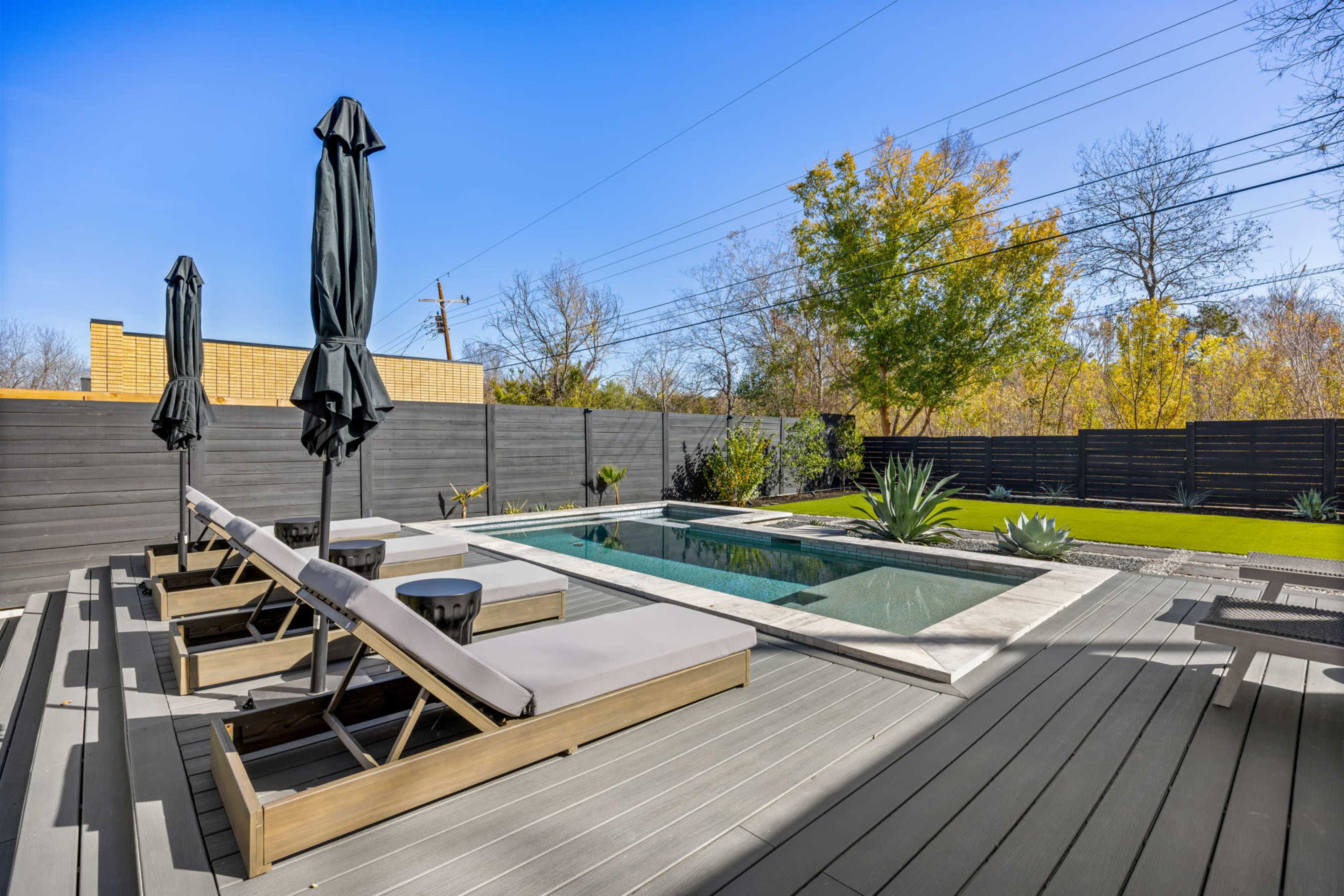 The image shows a modern backyard featuring a small swimming pool surrounded by lounge chairs and outdoor umbrellas, with a landscaped area and a wooden fence.