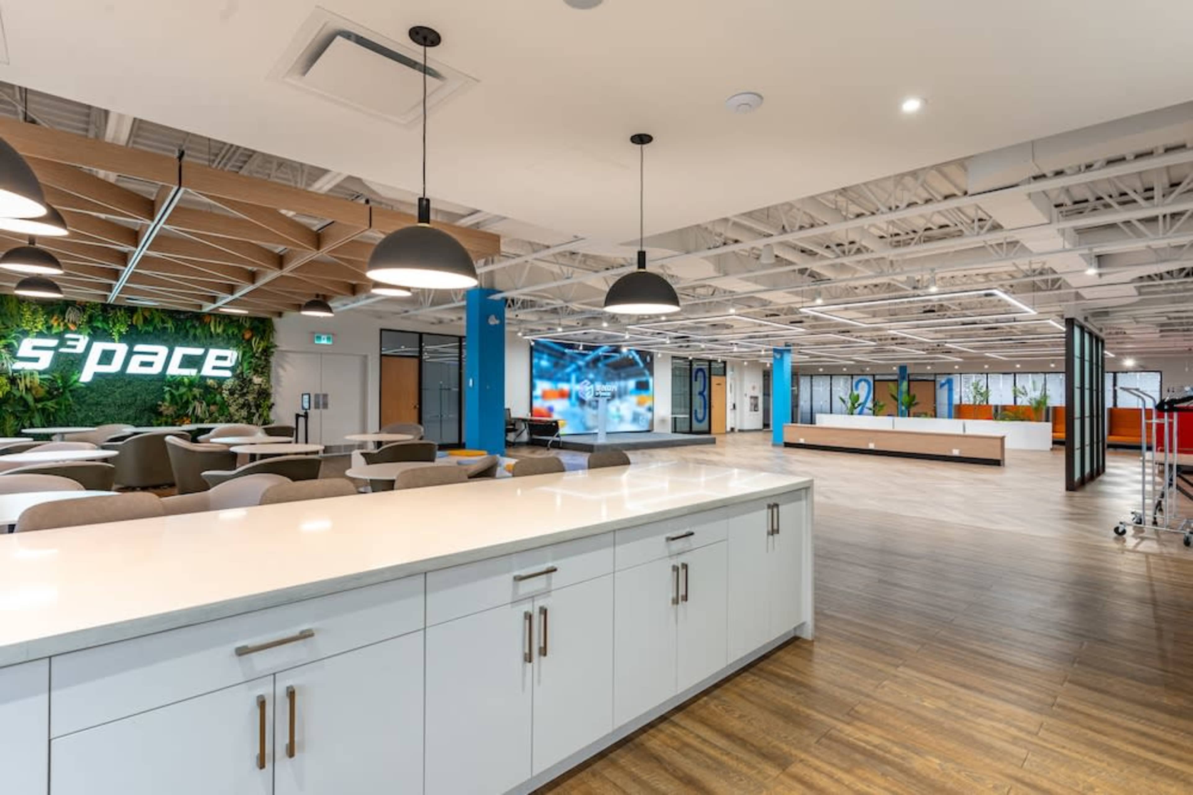 The image shows a modern office interior featuring a common seating area, a kitchen island, and large windows with greenery decor on one wall.