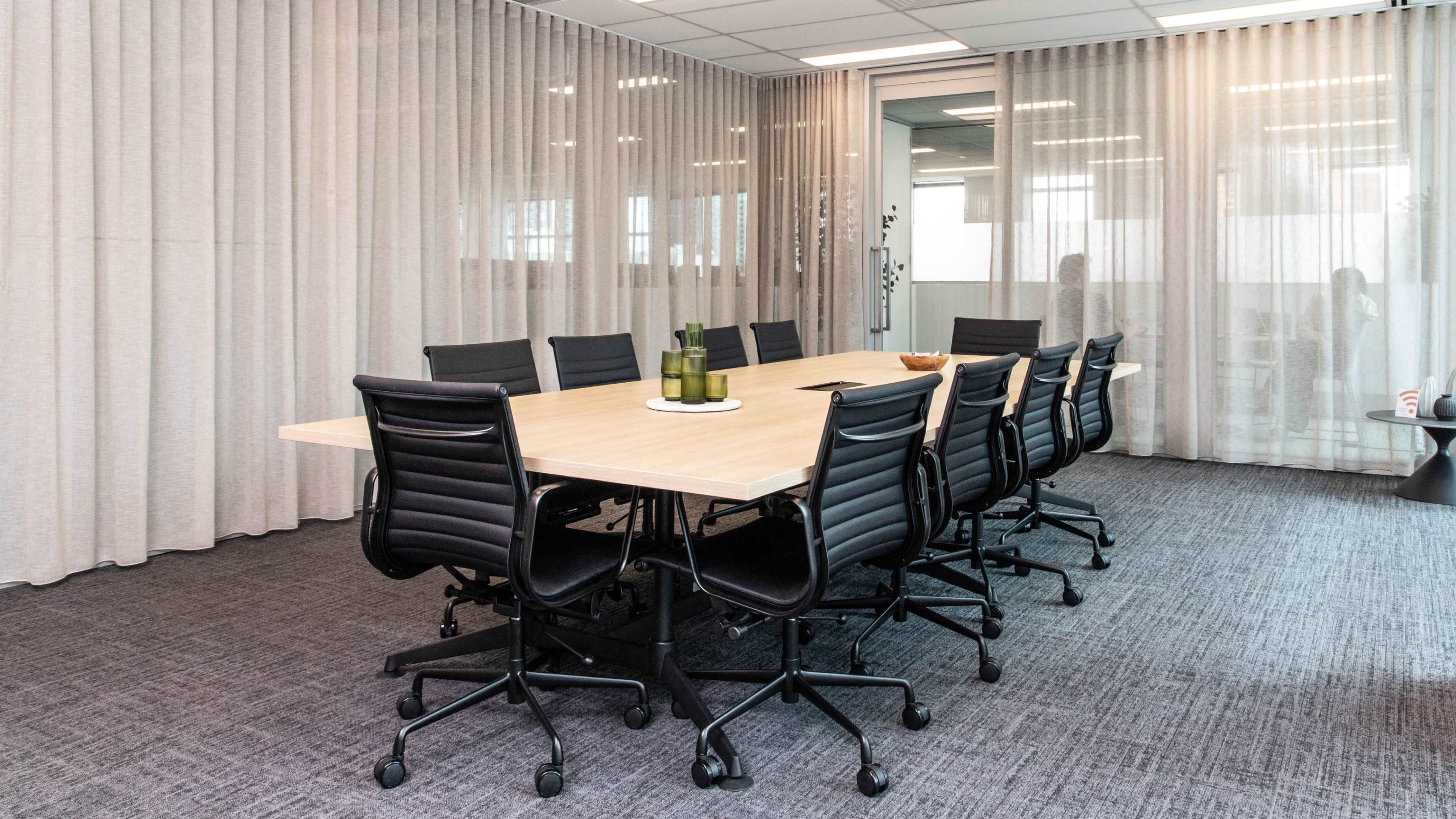 A modern conference room features a long wooden table surrounded by black rolling office chairs, with sheer curtains dividing the space.
