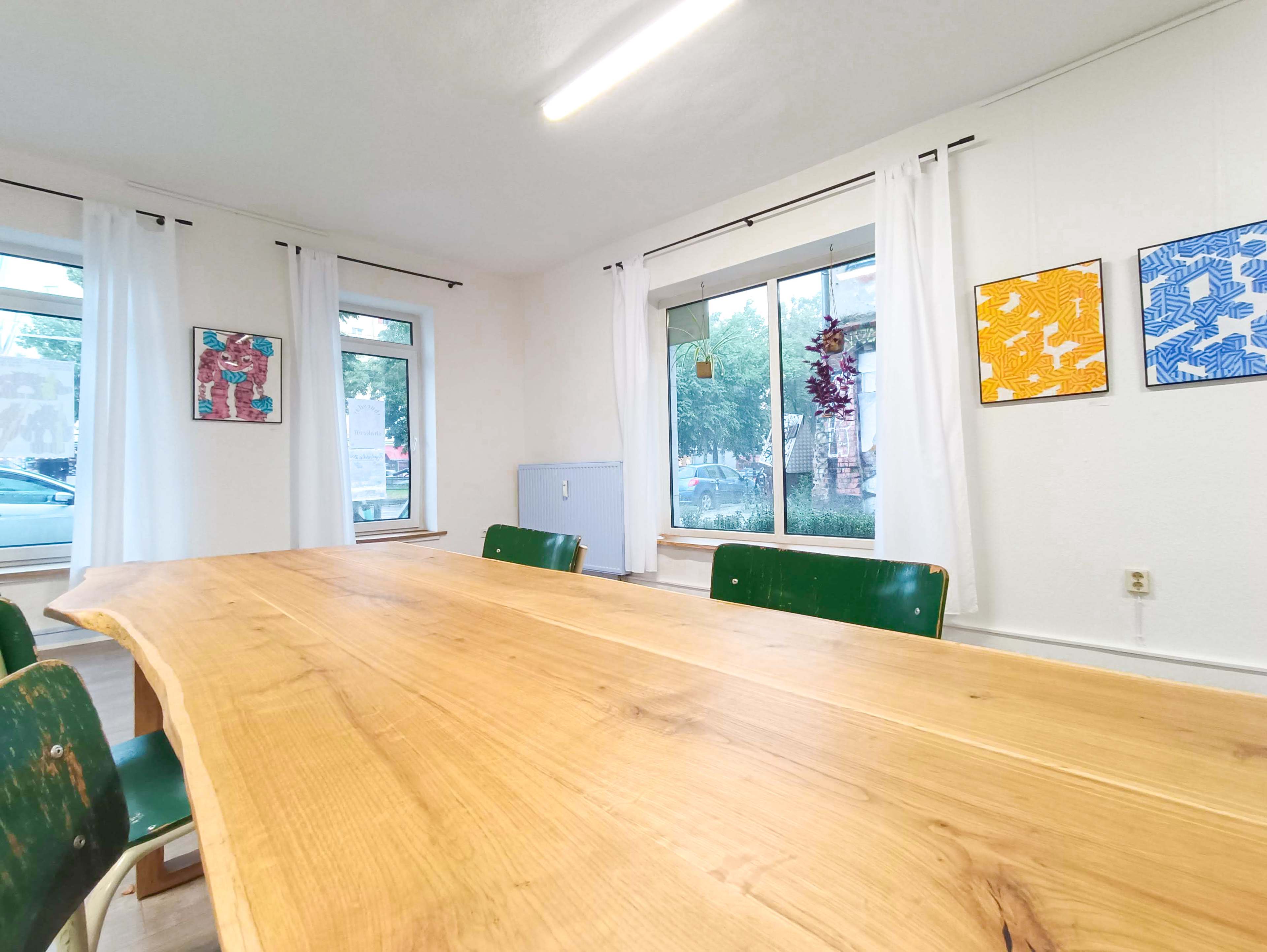 The image shows a wooden dining table with green chairs, set in a room featuring large windows and framed artwork on the walls.
