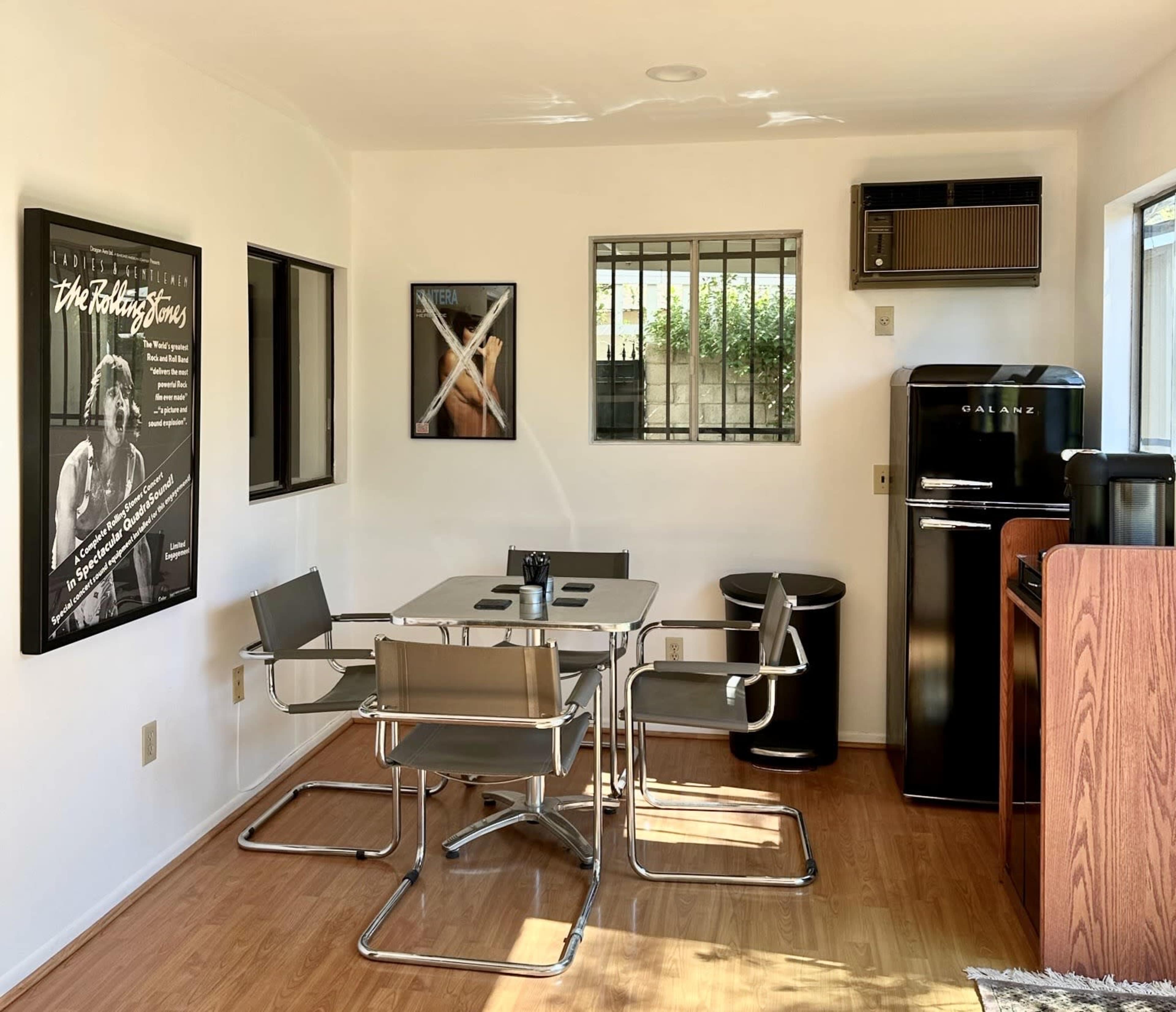 A small dining area with a glass table and metal chairs, accompanied by a refrigerator, waste bin, and framed posters on the walls.