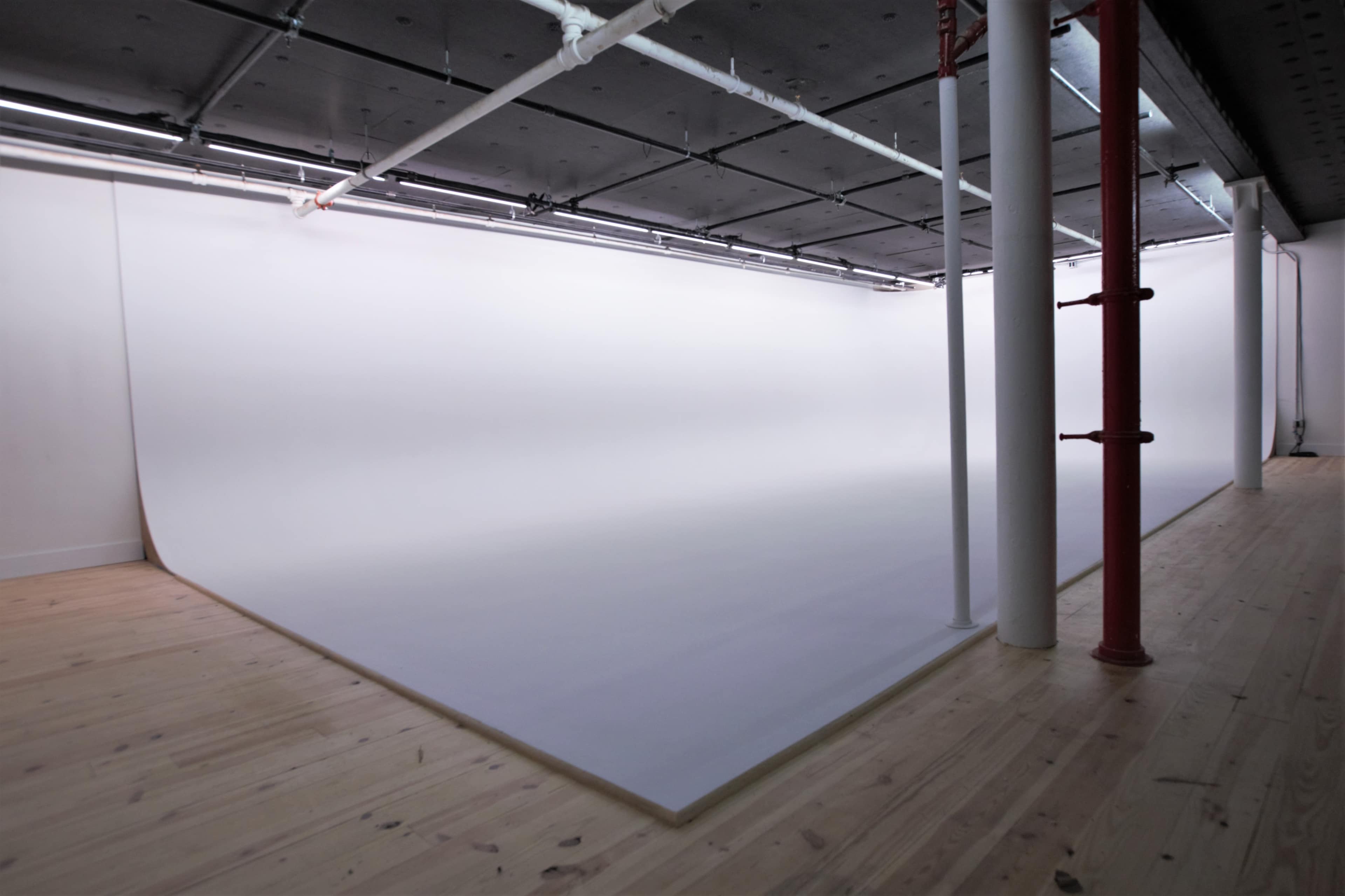 The image shows a minimalist photography studio with a large white backdrop and wooden flooring.