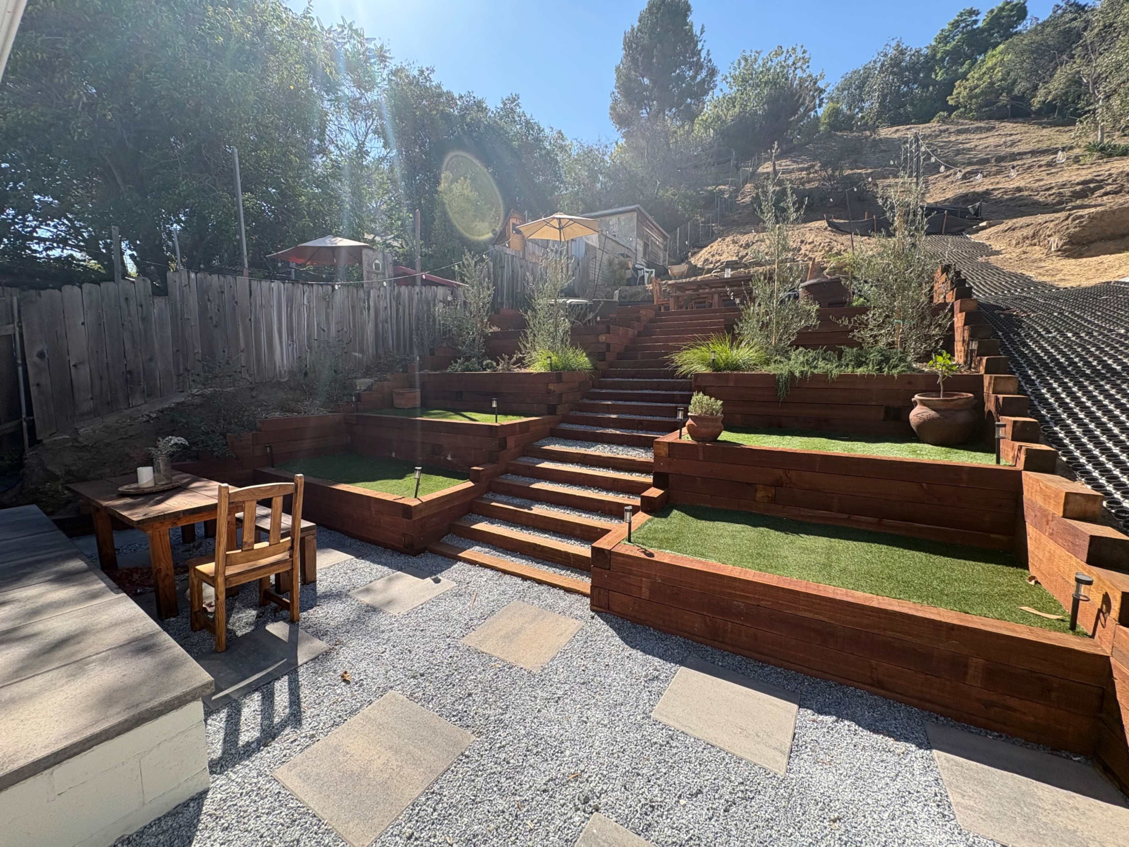 The image shows a landscaped backyard with terraced wooden planters, stone steps leading up, and a table and chairs in a gravel area.